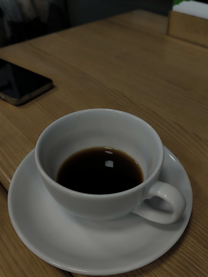 A white ceramic cup, partially filled with a dark liquid resembling coffee, rests on a matching white ceramic saucer. The cup features a handle on its right side. Both items are positioned on a light brown wooden table surface, exhibiting visible wood grain. To the upper left of the cup, a dark-colored smartphone lies flat on the table. In the upper right background, a wooden container holds several white rectangular items, likely napkins, with a subtle green element visible behind them. The lighting suggests an indoor setting. The specific location within Vinnytsia, Ukraine, cannot be determined from the visual evidence provided in the image. No individuals are present or discernable, and no actions or interactions are depicted.