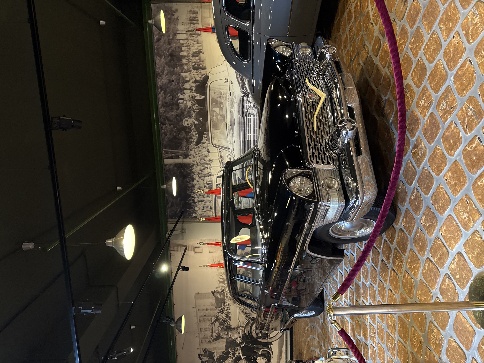 A black GAZ-13 Chaika limousine, manufactured in the Soviet Union, is displayed indoors in front of a large historical mural depicting a public event with crowds and prominent figures. The car is in pristine condition, with a chrome grille and yellow emblem. The interior, visible through the windows, appears to have red seating. The car is roped off with velvet stanchions. The floor is paved with decorative square tiles. Several industrial-style pendant lights hang from the dark ceiling, illuminating the scene. The mural behind the car shows two men in formal attire, one waving, standing in an open-top vehicle, with a crowd of people in the background. Another vehicle, a grey sedan, is partially visible to the right of the black car. The overall setting suggests a museum exhibit in the city of Arkhangel’skoye, Russia, possibly dedicated to Soviet-era history or automobiles.