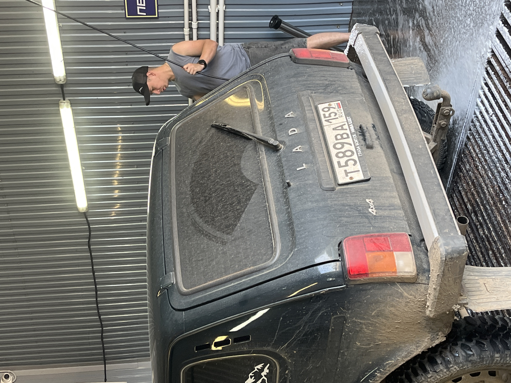 A man in a gray t-shirt and shorts, wearing a black baseball cap, stands to the right of a dirty, dark green Lada Niva car. He is holding a long pole, likely a car wash brush, and appears to be washing the rear of the vehicle in an outdoor car wash bay. The car has a visible license plate that reads "T589BA 159 RUS". The ground in front of the car is wet and foamy, indicating an active car wash. The bay has corrugated metal walls and fluorescent lighting fixtures overhead. The scene appears to be in Chaikovsky, Russia, as suggested by the car's license plate and the context provided.