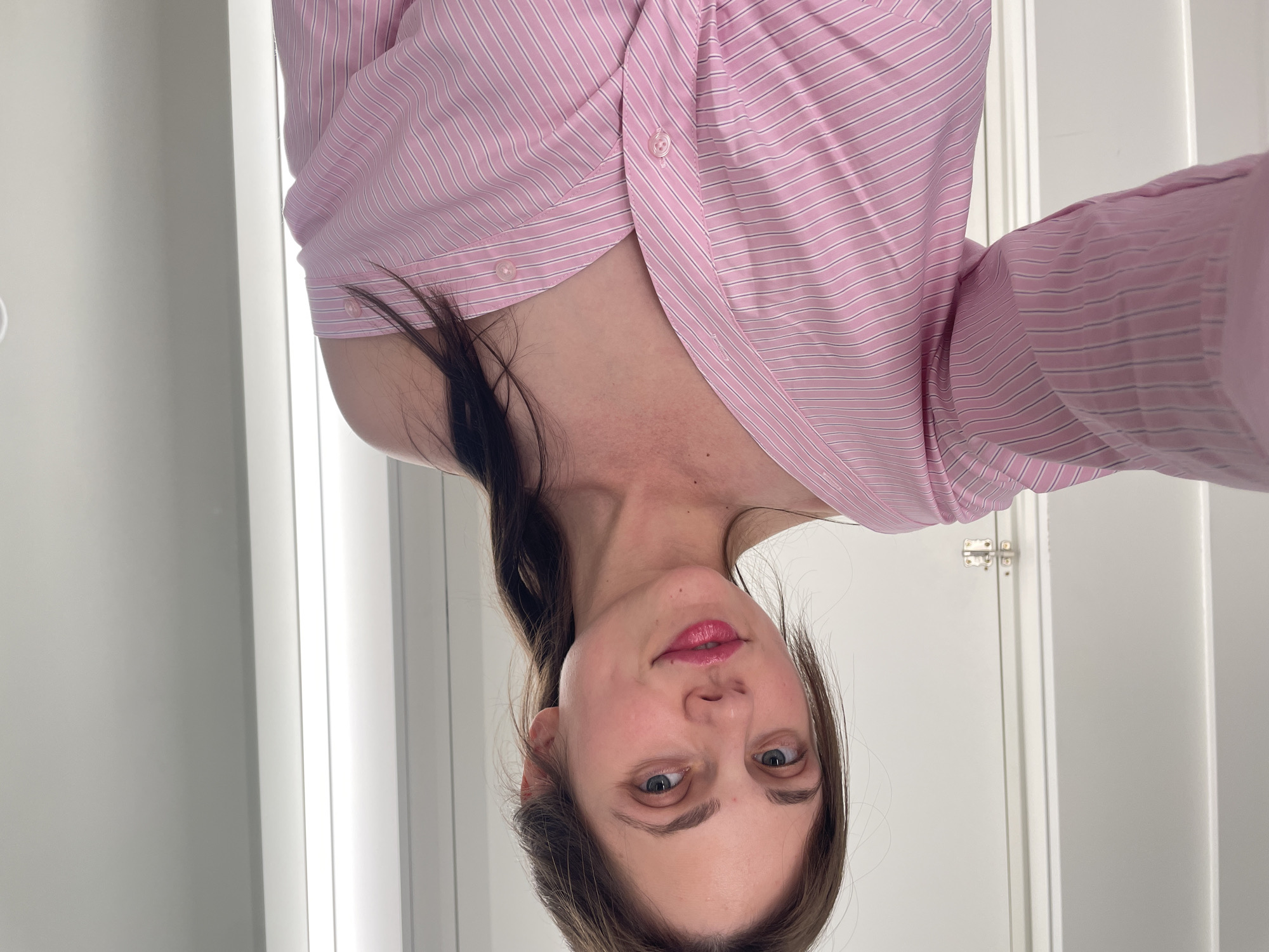 The image shows a woman with brown hair and blue eyes, wearing a pink and white striped off-the-shoulder shirt. She is looking at the camera and smiling. The background is a plain white wall with a vertical light fixture on the right side and a door on the left side. The scene appears to be indoors, possibly in a fitting room or a well-lit closet. There is no visible indication of the location being in Vecsés, Hungary.