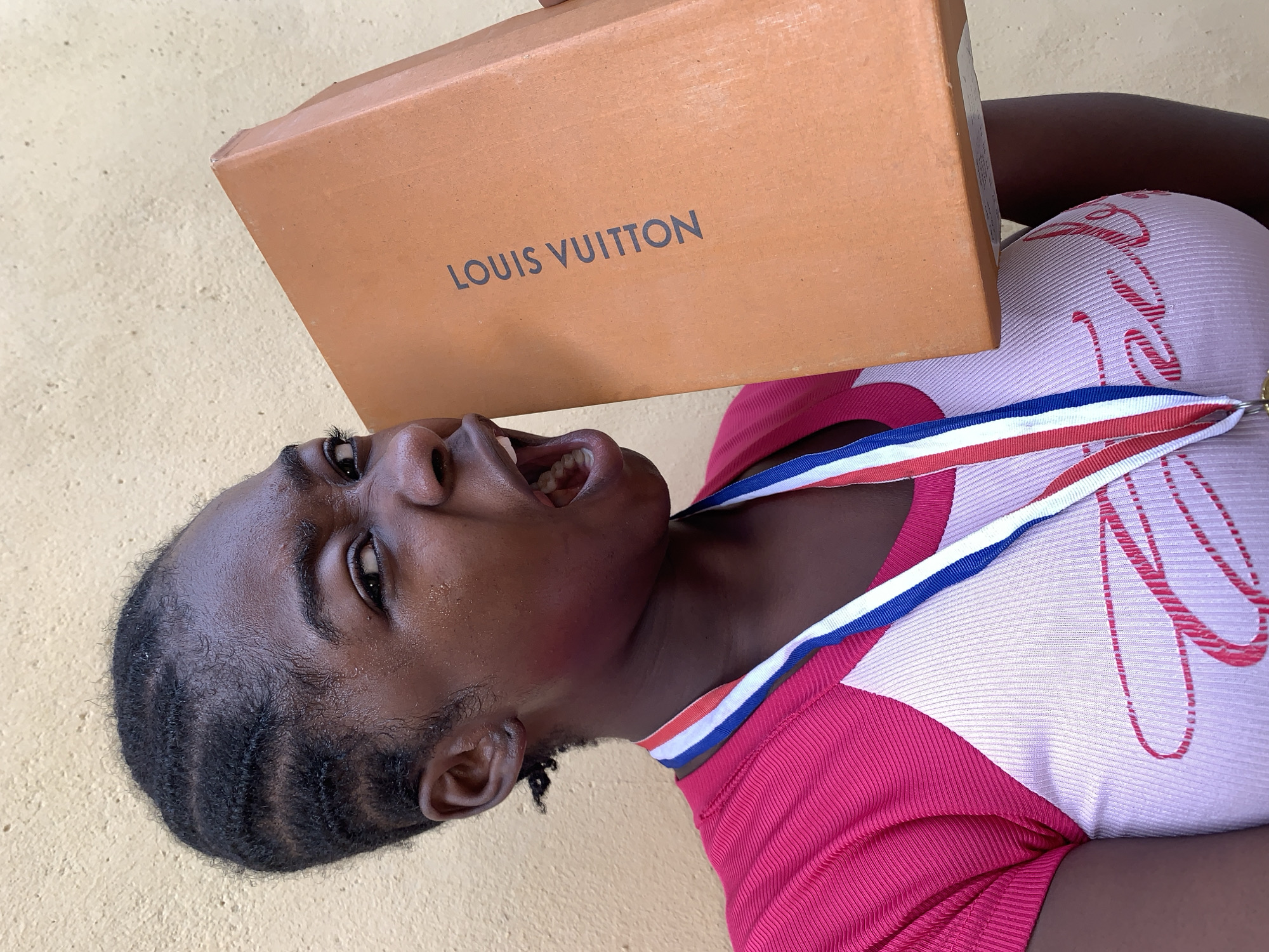 An individual is lying on a light yellow surface, possibly a floor or ground, holding a Louis Vuitton box above their head. The individual has dark skin, short braided hair, and is wearing a pink and white shirt. A red, white, and blue ribbon is draped around their neck. The person's mouth is open. The location is Rafin Par, Nigeria.