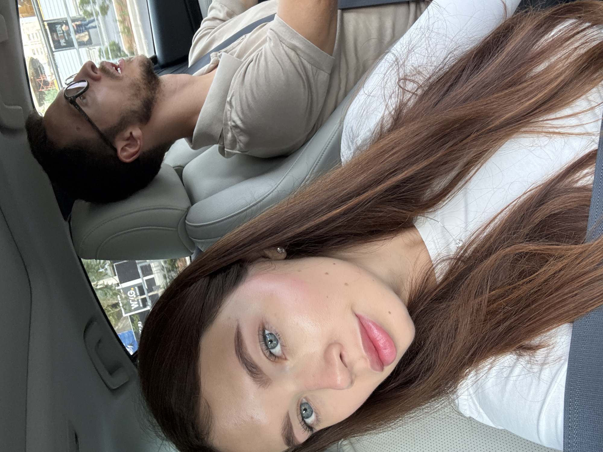 A woman with brown hair and blue eyes, wearing a white shirt, is sitting in the passenger seat of a car. She's looking at the camera. A man with dark hair and a beard, wearing glasses and a beige shirt, is in the driver's seat with his head reclined back on the headrest. The car interior is grey leather. Outside the car window, there are buildings and signage, including one with "WAG" and a word ending in "TEN," suggesting a location in a developed urban area, possibly Tel Aviv, Israel.