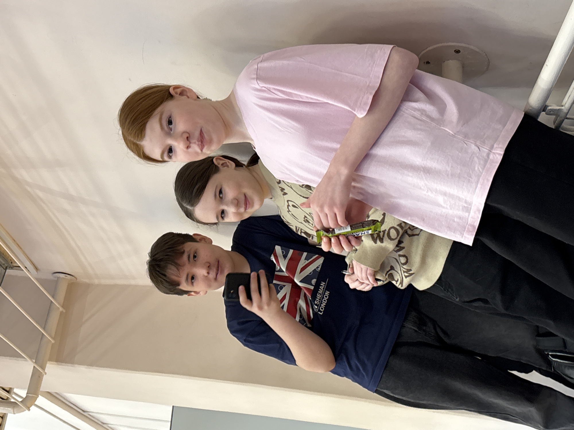 Three teenagers stand close together in what appears to be an indoor setting. The person in front, a boy, holds a smartphone, possibly taking a selfie. He wears a dark blue t-shirt with a British flag design. Behind him is a girl holding a packaged snack bar in her hands. She wears a light-colored patterned sweater. At the top, a girl with red hair wears a pink t-shirt. The background is a light-colored wall, partially revealing a window and a pipe structure. The location is Izhevsk, Russia.