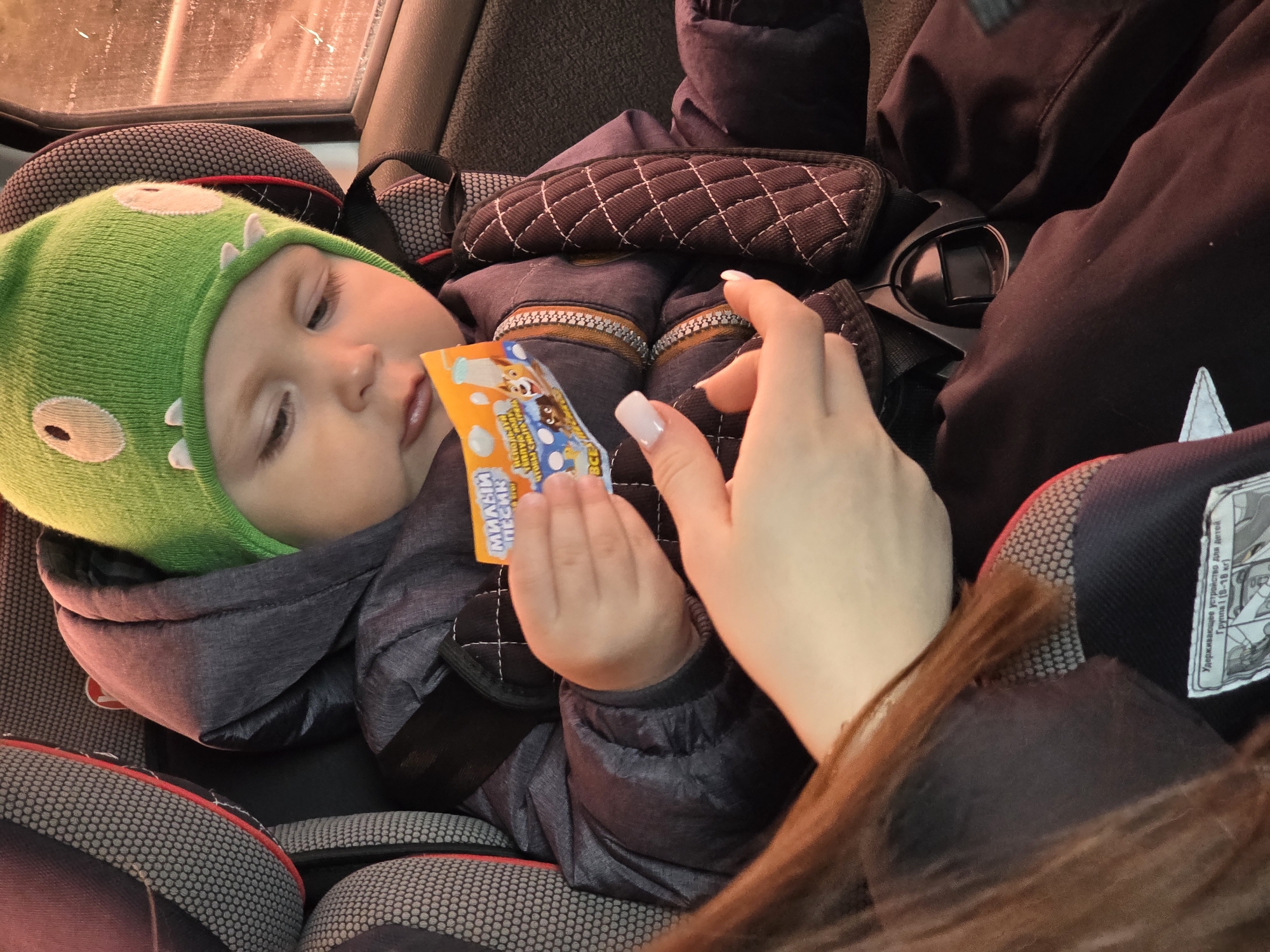 A baby wearing a green hat with cartoon eyes lies in a car seat. The baby holds a small, orange packet in their hand. A person's hand, with manicured nails, is positioned near the baby's hand. The car seat is black and gray. A portion of a black jacket is visible in the upper right of the image. The Russian text on the packet indicates "Милый ПЕСИК" ("Cute Doggy"), "Используй BCE" ("Use All"). Another Russian text says "Удерживающее устройство для детей Группа 1 (9-18 кг)" ("Restraint device for children Group 1 (9-18 kg)"). The image was taken in Chaikovsky, Russia.