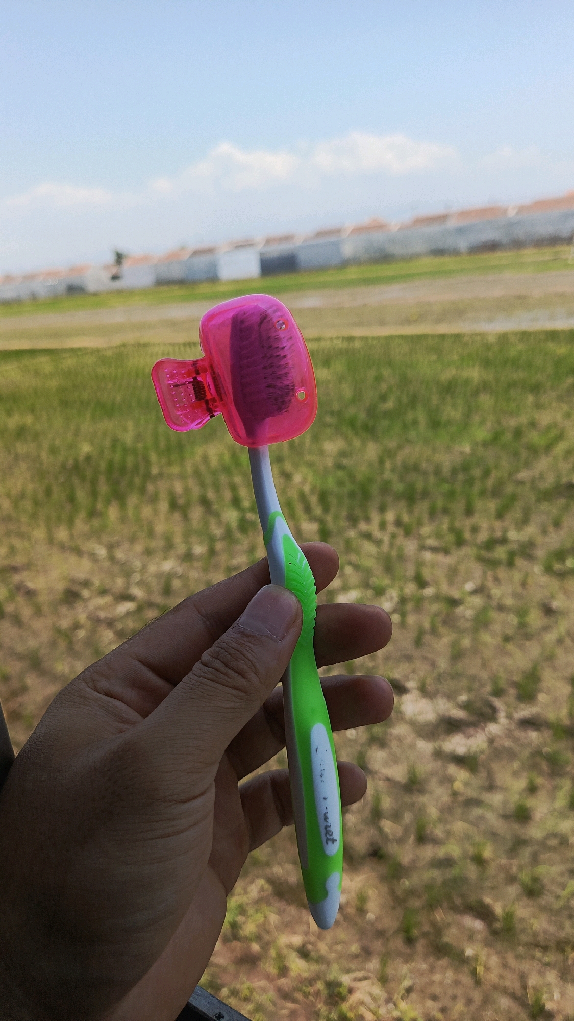 A person's left hand holds a toothbrush with a bright pink cap on its bristles. The toothbrush has a white handle with green accents and appears to be made of plastic. The background is a blurred landscape of green fields, a white wall, and a pale blue sky with scattered clouds. The scene suggests an outdoor setting, possibly in Babadan, Indonesia, given the vegetation and architecture visible. The focus is on the toothbrush and hand, with the background out of focus.