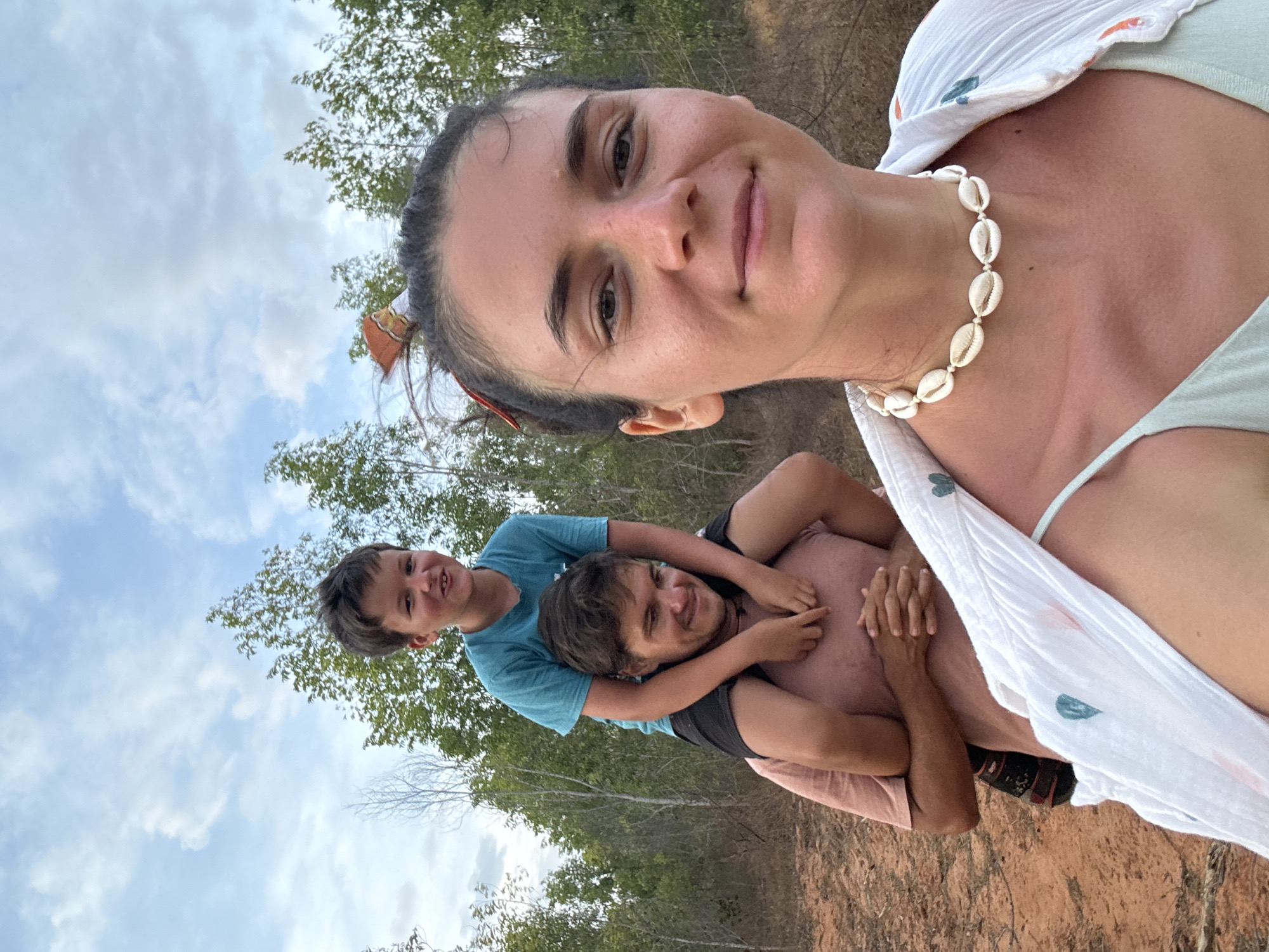 The image is a selfie of a woman with two males who are likely her children. The woman is fair-skinned with dark hair, wearing a seashell necklace and a white coverup over a light-colored top. One child is perched on the shoulders of the other male. The top child is wearing a turquoise shirt. The bottom male's face is visible; he is wearing a black shirt and pink shorts. In the background, there are trees, clouds, and a sandy ground, suggesting an outdoor location. Phu Thuy Ward, Vietnam is likely the location. The interaction is familial, with the individuals posing for a photo.