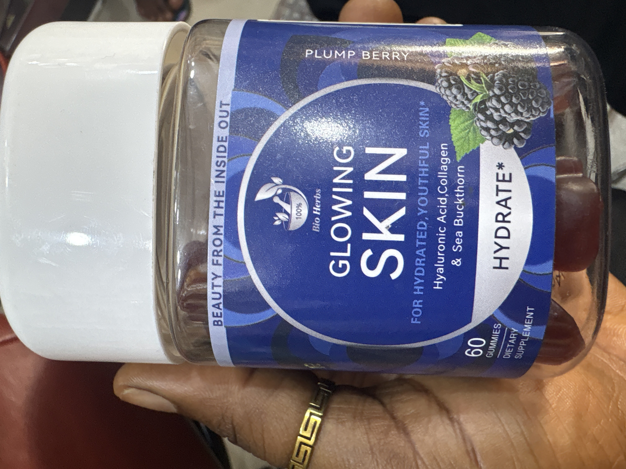 A person's hand, adorned with a gold ring, is holding a clear plastic bottle containing "GLOWING SKIN" gummies by Bio Herbs. The bottle has a white lid and a blue label featuring images of blackberries and text indicating "PLUMP BERRY" flavor and the ingredients "Hyaluronic Acid, Collagen & Sea Buckthorn." The label also states "60 GUMMIES DIETARY SUPPLEMENT" and "HYDRATE*". The background appears to be a blurred indoor environment with hints of red and black. There is no discernible connection to the city of Jos, Nigeria.