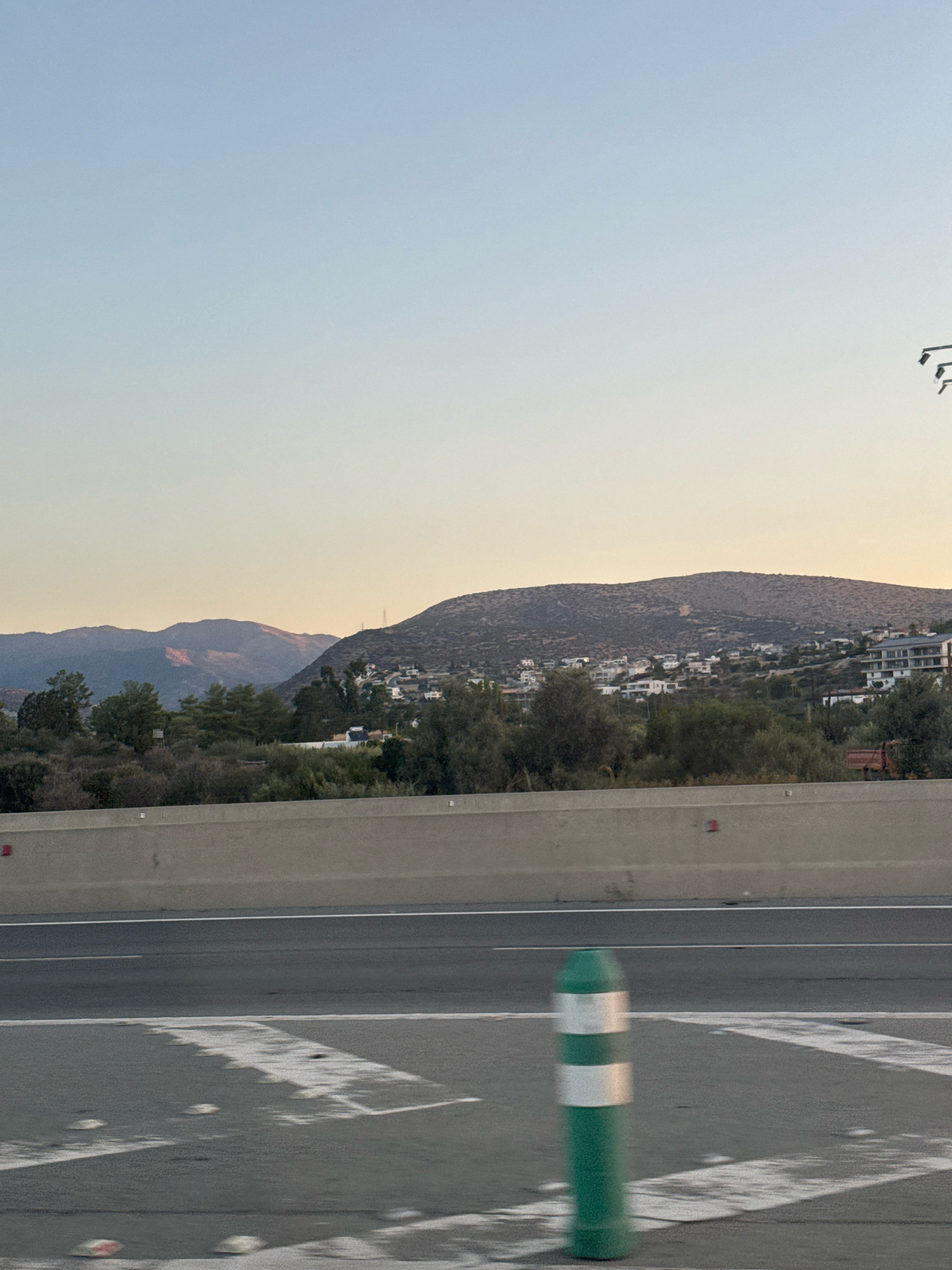 An outdoor, medium shot captures a road leading towards a hillside dotted with buildings under a clear, pale blue sky. The foreground shows a grey asphalt road with white painted markings and a green and white striped traffic bollard. A concrete barrier separates the road from a densely vegetated area. Beyond the vegetation, a village or town sprawls across the lower slopes of a large, undulating hill. The buildings are predominantly white with dark roofs, creating a contrast against the natural landscape. Further in the distance, a mountain range is visible, bathed in the soft light of what appears to be either dawn or dusk, with some peaks catching a warm, reddish-orange hue. The scene suggests a suburban or semi-rural setting within the Germasogeia Municipality of Cyprus, possibly captured from a vehicle in motion due to slight motion blur.