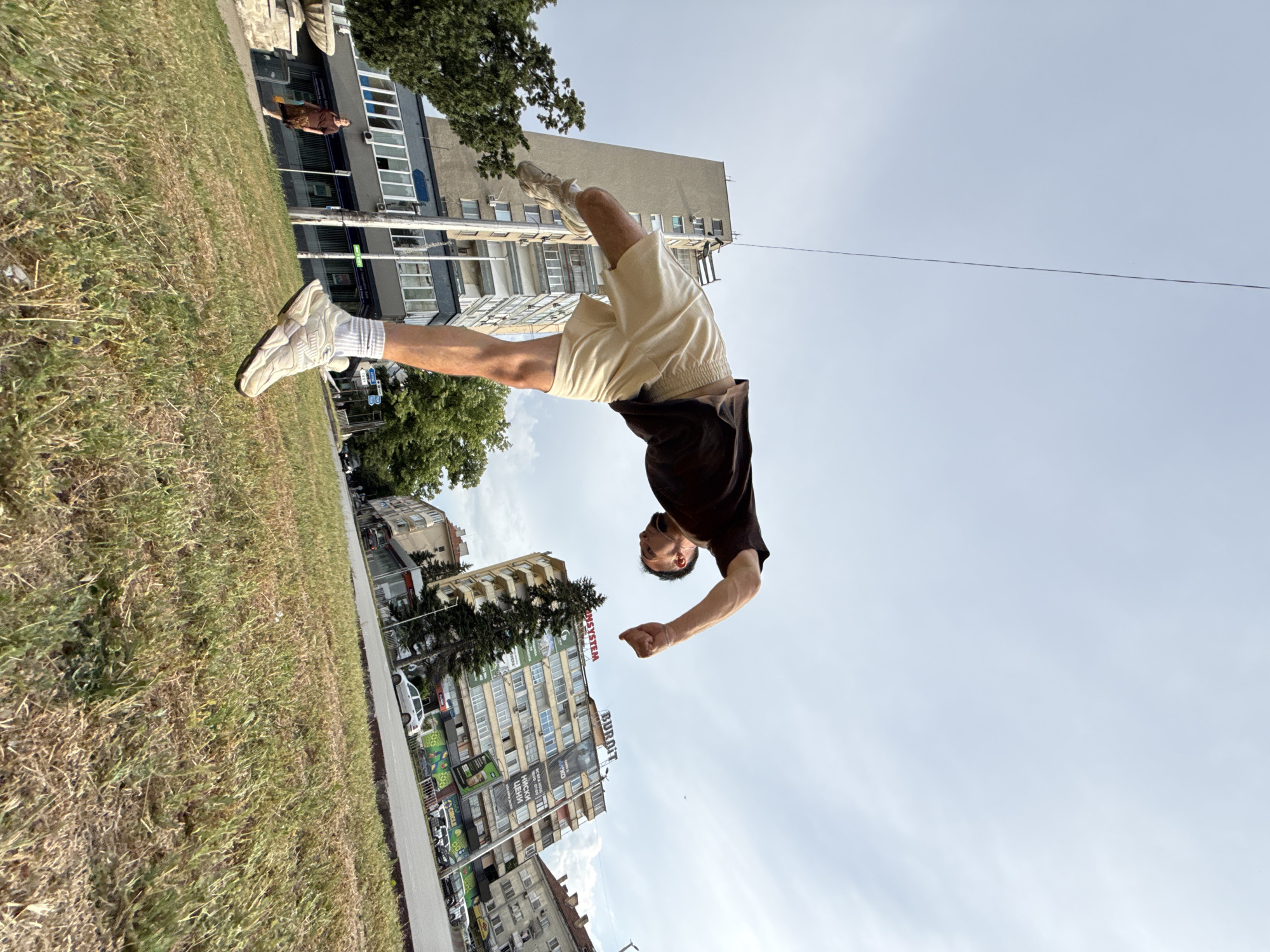 In Shumen, Bulgaria, a young man is captured mid-air performing a parkour move on a grassy incline with a cloudy sky above. He is wearing a dark t-shirt, light-colored shorts, white socks, and white athletic shoes. His body is bent backward, arms extended, with his right leg in front of him and his left leg extended behind. In the background, several multi-story buildings are visible, along with trees and a street with cars. A woman walks on a paved path to the left of the frame, and to the right, a sign for "BURBIT" is prominent on one of the buildings. The composition is wide-angle, emphasizing the action of the parkour athlete against the urban backdrop.