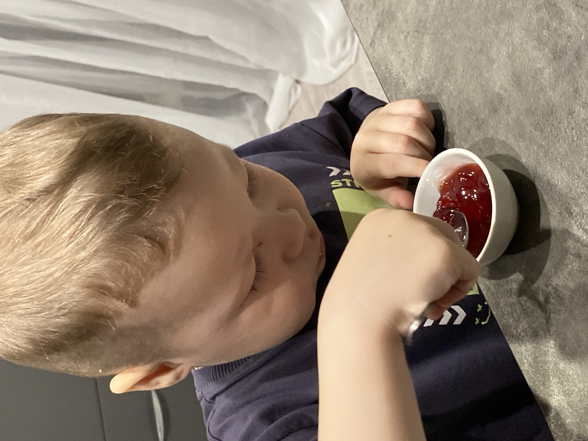 A high-angle, portrait-oriented photograph captures a young child with light blond hair at a gray, textured countertop. The child, whose face is partially visible in profile, wears a dark blue long-sleeved shirt. Their left arm and hand are visible, holding a small white spoon in a small white bowl filled with a glistening red jam-like substance, likely raspberry or strawberry jam. The child appears focused on the food, with their mouth slightly open, suggesting they are in the process of eating or about to take a bite.
On the child's dark blue shirt, a partial green graphic is visible, which includes white chevron arrows (`>>`), the letters "ST," and a small smiley face icon (`:`). In the background, a light-colored, possibly white, curtain or draped fabric is present, creating a soft, blurred backdrop. The lighting is even, suggesting an indoor setting. The scene conveys a moment of quiet concentration and enjoyment during a meal or snack time. The location context provided is Sochi, Russia.