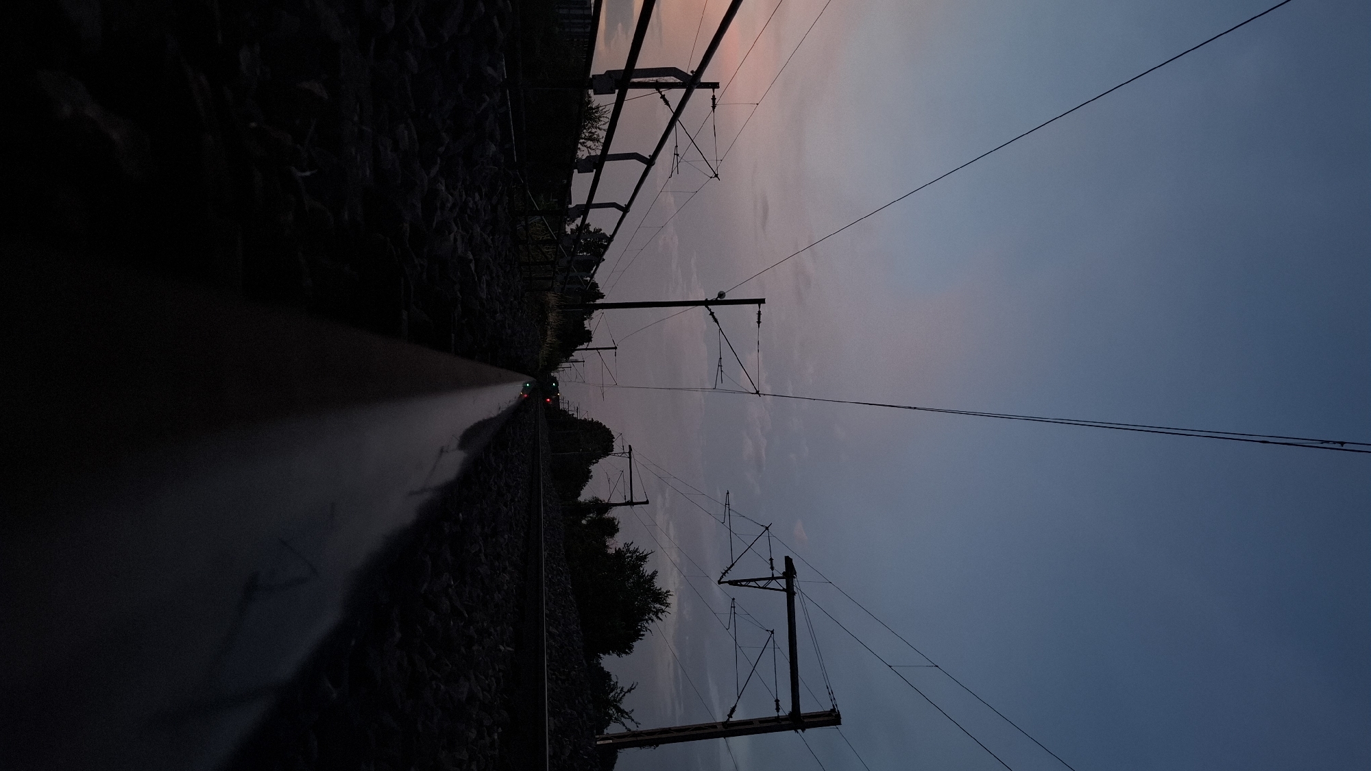 The image depicts a low-angle shot of a railway track at dusk, likely in Strasbourg, France, given the context. The sky is a gradient of deep blue at the top transitioning to lighter blue and hints of pink and purple near the horizon. Overhead, power lines for the electric trains stretch across the frame, supported by dark poles. The tracks themselves are made of ballast and two parallel rails, with one rail prominently in the foreground, reflecting the twilight sky and appearing wet or polished. To the left, a dark metal fence runs alongside the tracks, and beyond it, silhouetted trees and a building are faintly visible. On the right, more trees and vegetation are visible. In the distance, faint lights, possibly from an approaching train, are visible. The overall atmosphere is serene and slightly melancholic, typical of twilight in an urban or suburban environment.