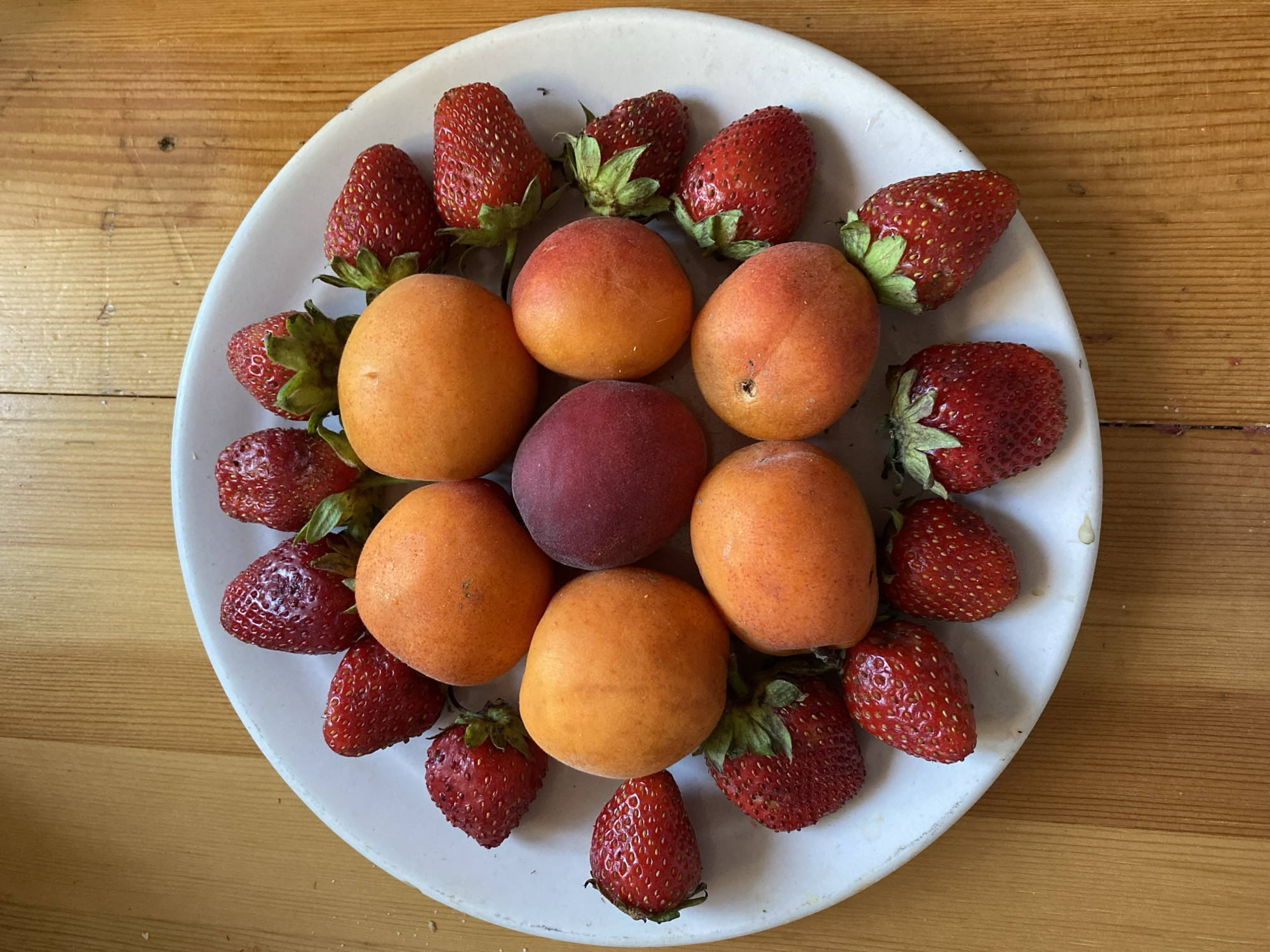 A close-up, overhead shot shows a white plate filled with fresh strawberries and apricots arranged on a wooden table. The plate is slightly off-center to the left, with the wooden table filling the right and bottom of the frame. The fruit is artfully arranged in a circular pattern around a single dark plum in the center. The strawberries are bright red, with green stems, and appear ripe and juicy. The apricots are a warm orange color, with a fuzzy texture, and are also plump and inviting. The wooden table has a natural grain and appears to be made of light-colored wood. The overall impression is one of freshness and abundance, with the vibrant colors of the fruit standing out against the rustic backdrop of the table.