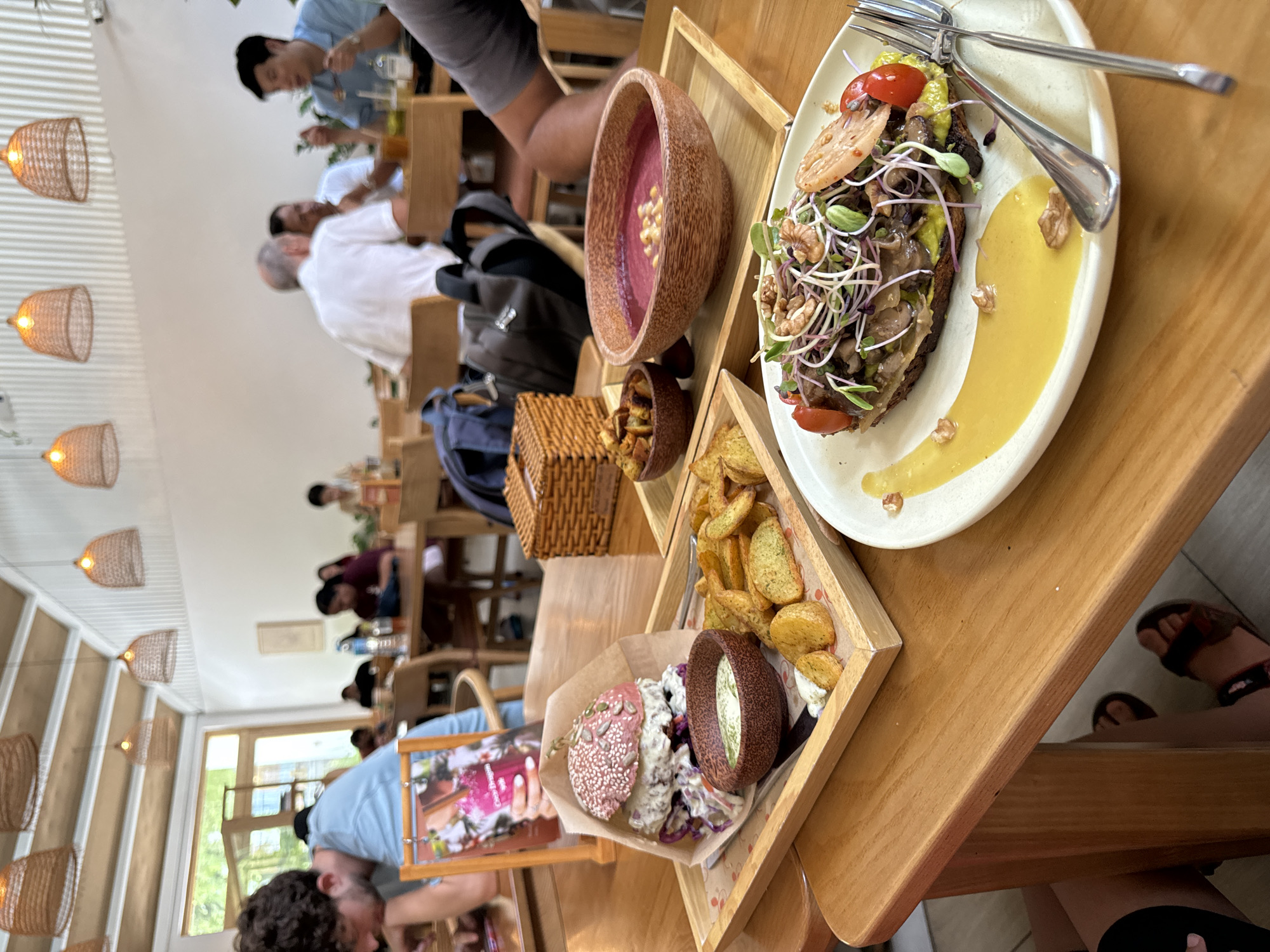 The scene depicts a restaurant in Thủ Đức, Vietnam. Several people are seated at tables, engaged in conversation or eating. The camera focuses on a table with a variety of food items: a dish with a bread-based item topped with vegetables and a yellow sauce, a bowl of reddish-purple soup with nuts, a small bowl of what appears to be fried croutons, a tray of potato wedges, and another tray with a burger featuring a pink bun and a bowl of coleslaw. The table is made of wood. Silverware is visible on one of the plates. Wicker lamps hang from the ceiling.