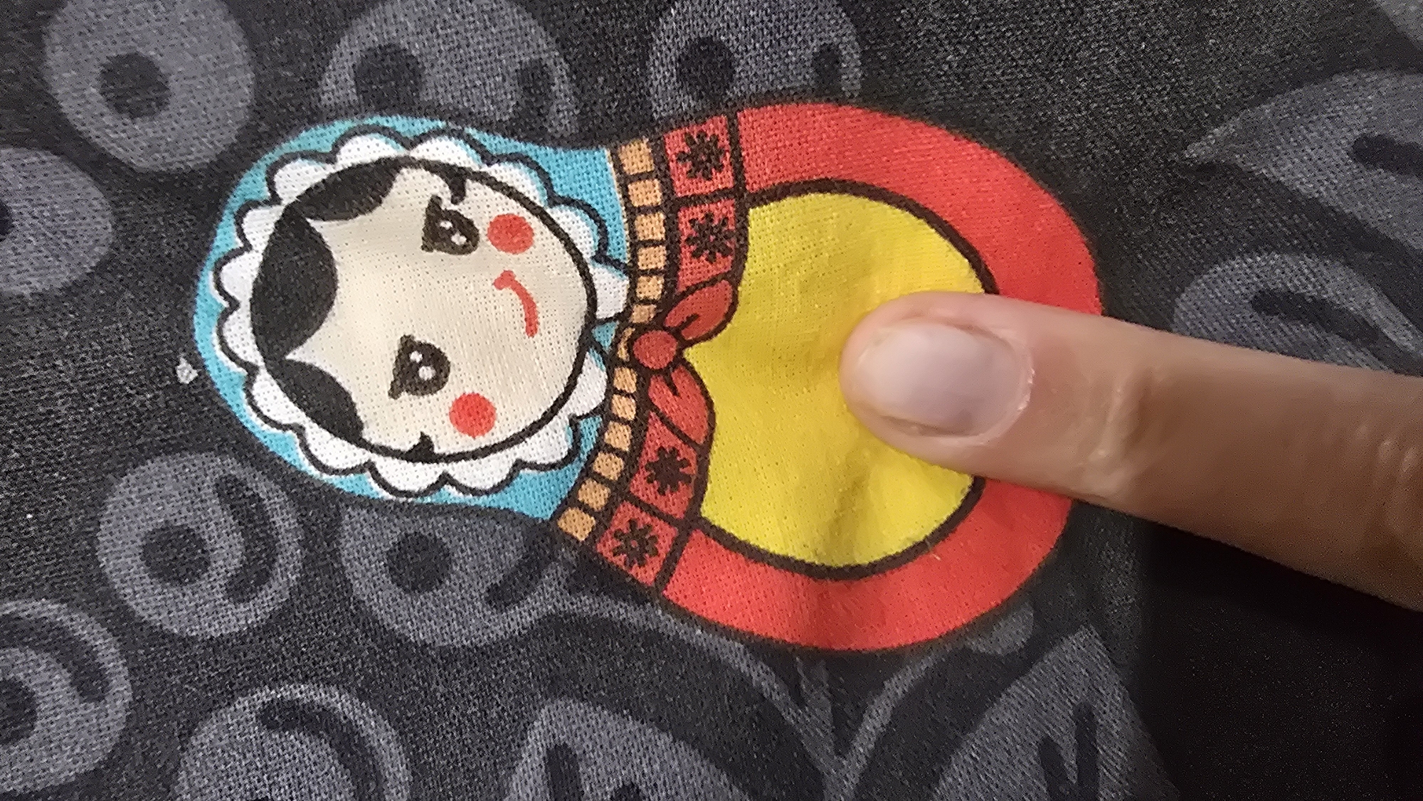 A close-up shot shows a finger pressing against a detail on a dark gray garment. The garment is printed with a pattern of faded smiley faces. In the foreground, a matryoshka doll illustration is prominently displayed. The doll has a light-colored face with rosy cheeks, dark eyes, and a smile. It wears a blue head covering trimmed with white scalloped lace. The doll's body is depicted in red and yellow, with decorative black and red patterns on its upper torso. The finger, with a visible fingernail, is obscuring a portion of the doll's yellow midsection. The image lacks any identifiable context of a specific location within the city of Chaikovsky, Russia, or any other city.