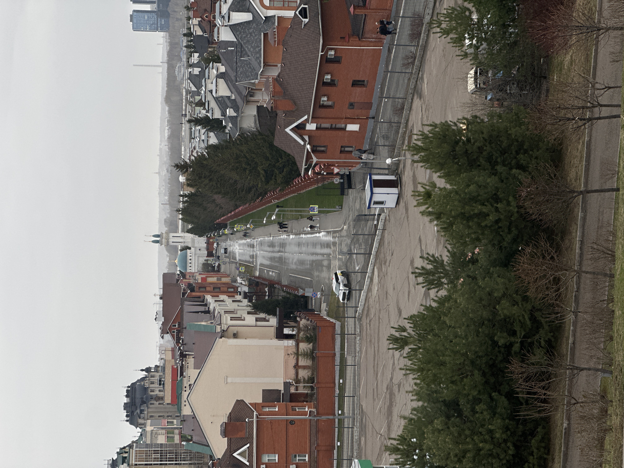 The image shows a view of the city of Kazan, Russia, on an overcast day. The buildings, mostly red and beige brick structures, line the wet street. A red fence separates the street from green spaces with trees. A white guard booth is visible on the right side of the street. Cars drive on the street. Pedestrians walk on the sidewalks. In the distance, the skyline shows a church with blue domes and tall buildings shrouded in haze. A parking lot is visible on the right.
