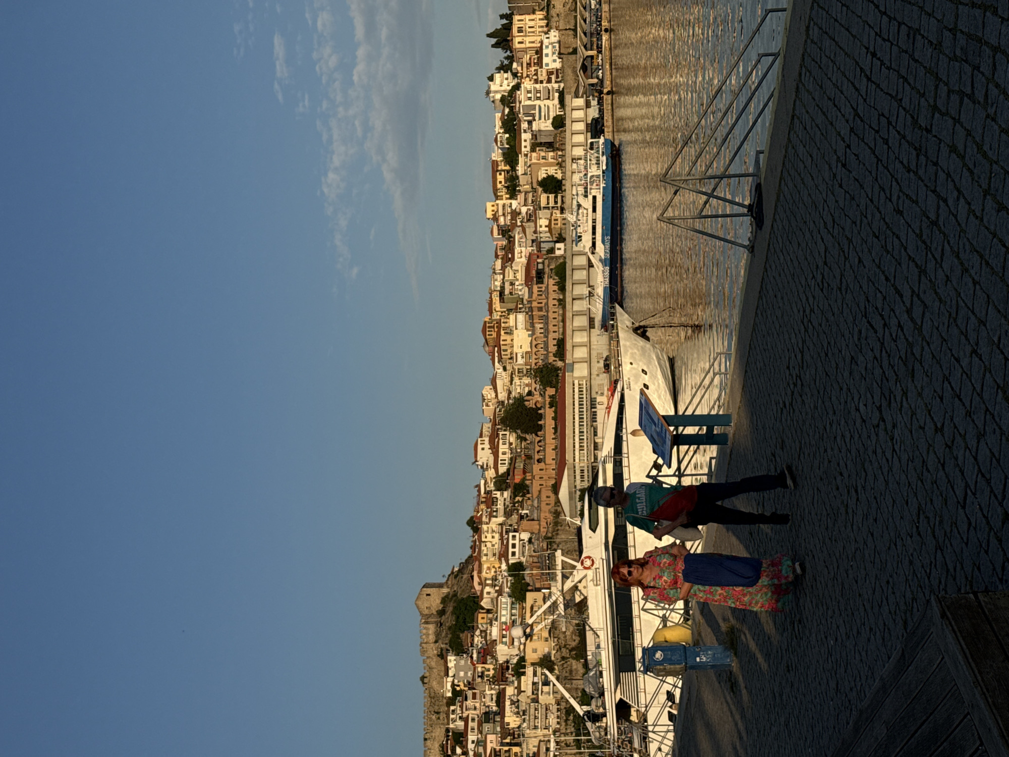 Here is a description of the image:
The image shows a view of Kavala, Greece, from a waterfront location. The photograph is rotated 90 degrees counter-clockwise. A steep hillside covered with buildings dominates the background. The buildings are mostly light-colored and vary in size and shape. The sky is blue with some clouds. In the foreground, there is a paved area, likely a sidewalk or pier. Two people are standing on the pavement. One person is wearing a colorful floral dress and sunglasses. The other is wearing a green shirt, dark pants, and sunglasses. Behind them, there are white boats docked along the waterfront. Metal railings are visible near the edge of the pavement. The water reflects sunlight.
