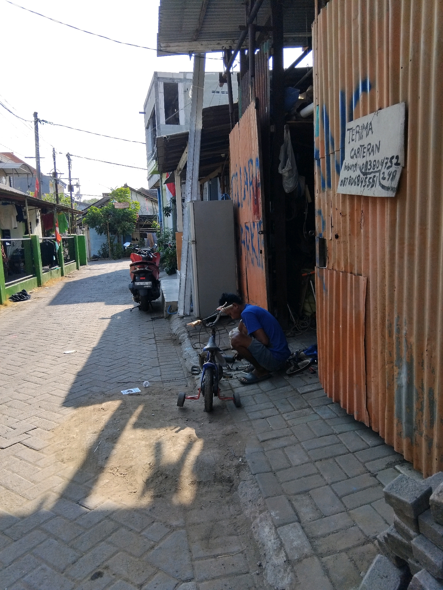 In the city of Kepuh Kiriman, Indonesia, on a cobblestone street lined with low-rise buildings, a man in a blue t-shirt and grey shorts kneels beside a small blue bicycle. He is engaged in working on the bicycle, which has training wheels. To his right, a corrugated orange metal structure forms the wall of a shop or workshop. A sign with handwritten text is affixed to the metal wall, reading "TERIMA CARTERAN" and providing contact numbers. A red scooter is parked further down the street. The scene is illuminated by sunlight, casting shadows across the street.