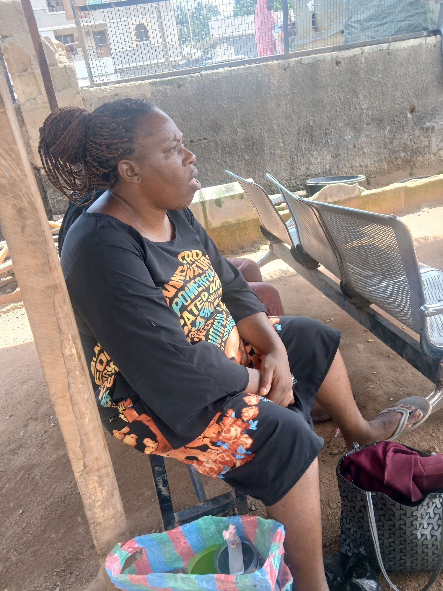 A woman with braided hair pulled into a bun sits on a metal chair in an outdoor waiting area. She is wearing a black long-sleeved shirt with a floral pattern on the bottom half and a colorful floral skirt. Her right hand rests on her left thigh, and her left hand is also on her lap. She is looking to her right with her mouth slightly open. In front of her are two metal benches. To her left, a wooden beam stands vertically. On the ground to her right is a dark grey handbag with a geometric pattern, and next to it, a plastic bag with colorful stripes and a green container inside. To her left, a multi-colored plastic bag with a green bowl is visible. The ground appears to be dirt. The background consists of a concrete wall and a metal fence, with some buildings visible beyond the fence.