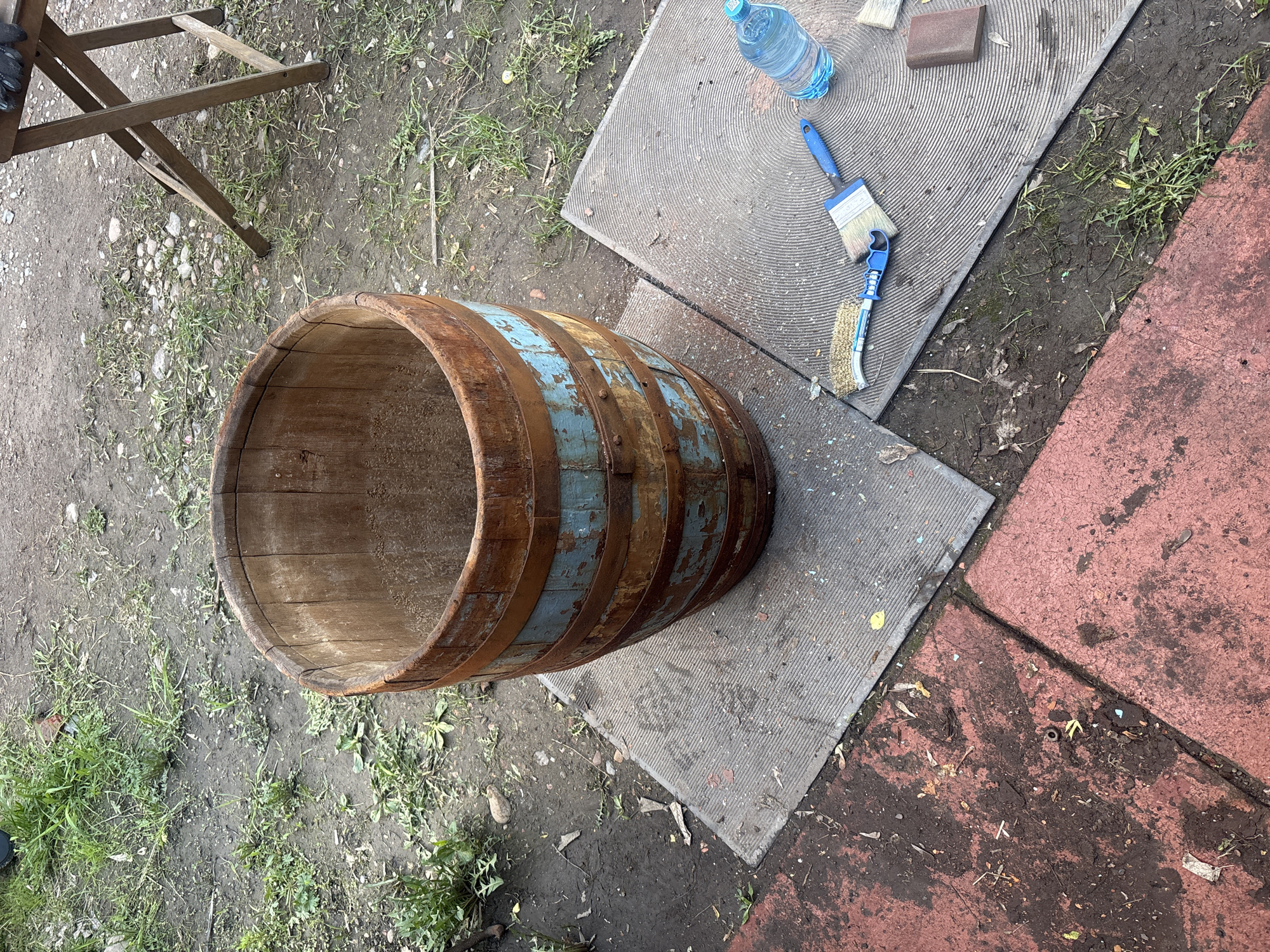 The scene is outdoors, likely in a yard or garden, potentially in Krasnoyarsk, Russia. The primary subject is a wooden barrel, possibly repurposed as a planter, with rusted metal bands and remnants of blue paint. The barrel sits partially on a gray, patterned concrete paving stone and partially on dirt and grass. Adjacent to the paving stone is a section of reddish-brown paving stones.
On the gray paving stone, near the barrel, are various objects: a blue water bottle, a sanding block, and two paintbrushes with blue handles. A section of a wooden stepladder is visible in the upper left corner. The ground is covered with dirt, grass, and small stones.