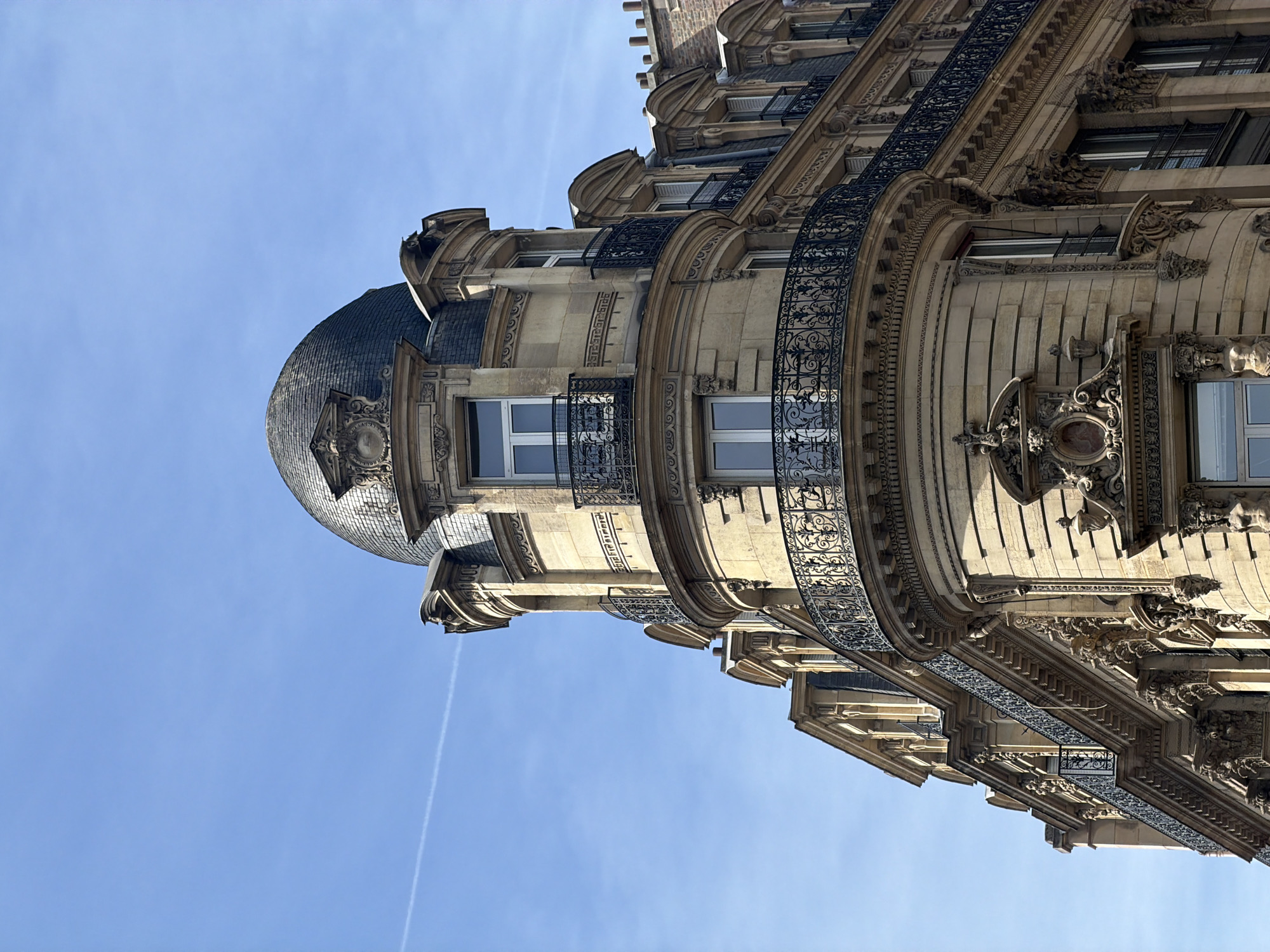 The image displays a detailed view of a grand, ornate building, typical of Parisian architecture, set against a clear blue sky. The photograph is taken from a low, upward-looking angle, with the building appearing rotated, highlighting its impressive height and design.
A prominent feature is a majestic domed or turreted roof section, covered in grey, scale-like tiles, possibly slate or zinc. Below this, and along the building's curved facade, are several balconies. These balconies are characterized by intricate black wrought-iron railings, featuring elegant scrollwork and floral patterns.
The building's facade is constructed from a light-colored stone, likely limestone or sandstone, and is lavishly adorned with detailed carvings, decorative reliefs, and pronounced cornices. Sunlight bathes the structure, indicating a bright daytime setting with a clear sky, marked only by a faint white contrail from an aircraft. Simple white-framed windows are visible between the ornate elements.
No people or activities are depicted; the focus is entirely on the architectural splendor. The scene conveys a sense of historical elegance and grandeur in an urban environment. There is no visible text in the image.