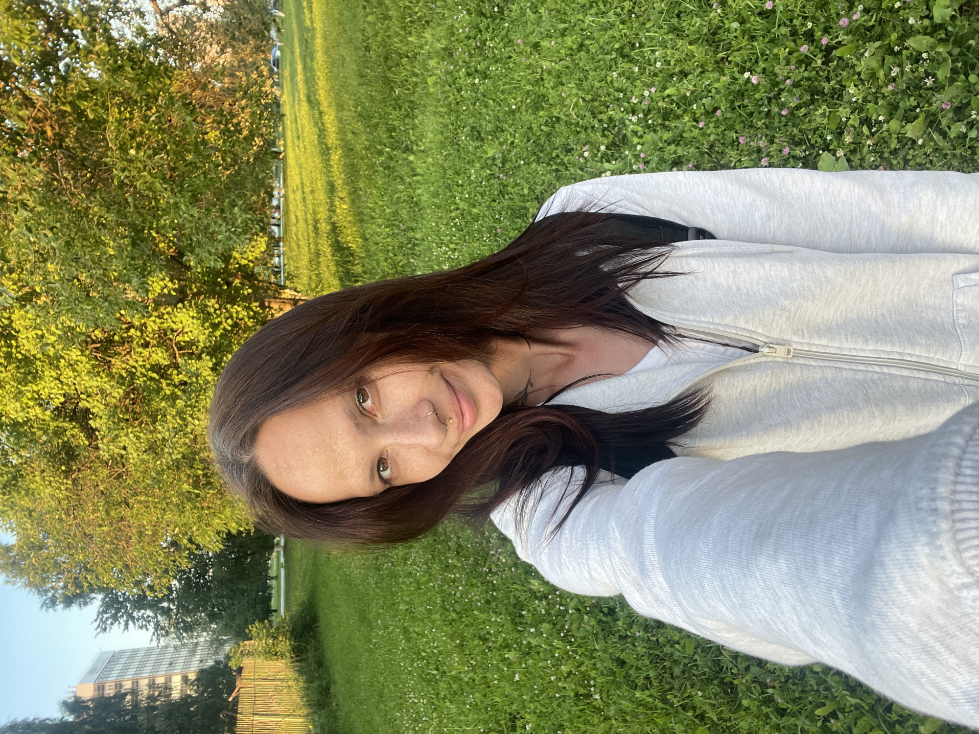 A young woman with long, dark brown hair and brown eyes smiles at the camera. She is wearing a light grey zip-up hoodie. Her face is tilted slightly to the left, and she has a nose ring and a small mole near her lip. Freckles are visible across her cheeks and nose. The background is a grassy field with small white and pink wildflowers. Sunlight casts long shadows, indicating it is late afternoon or early evening. To the left of the frame, a large tree with green leaves is visible, with a tall, modern building in the distance. The overall setting appears to be a park or open green space. The image has been taken from a low angle, possibly by the subject herself. No visible text is present in the image. The implied location is Izhevsk, Russia.