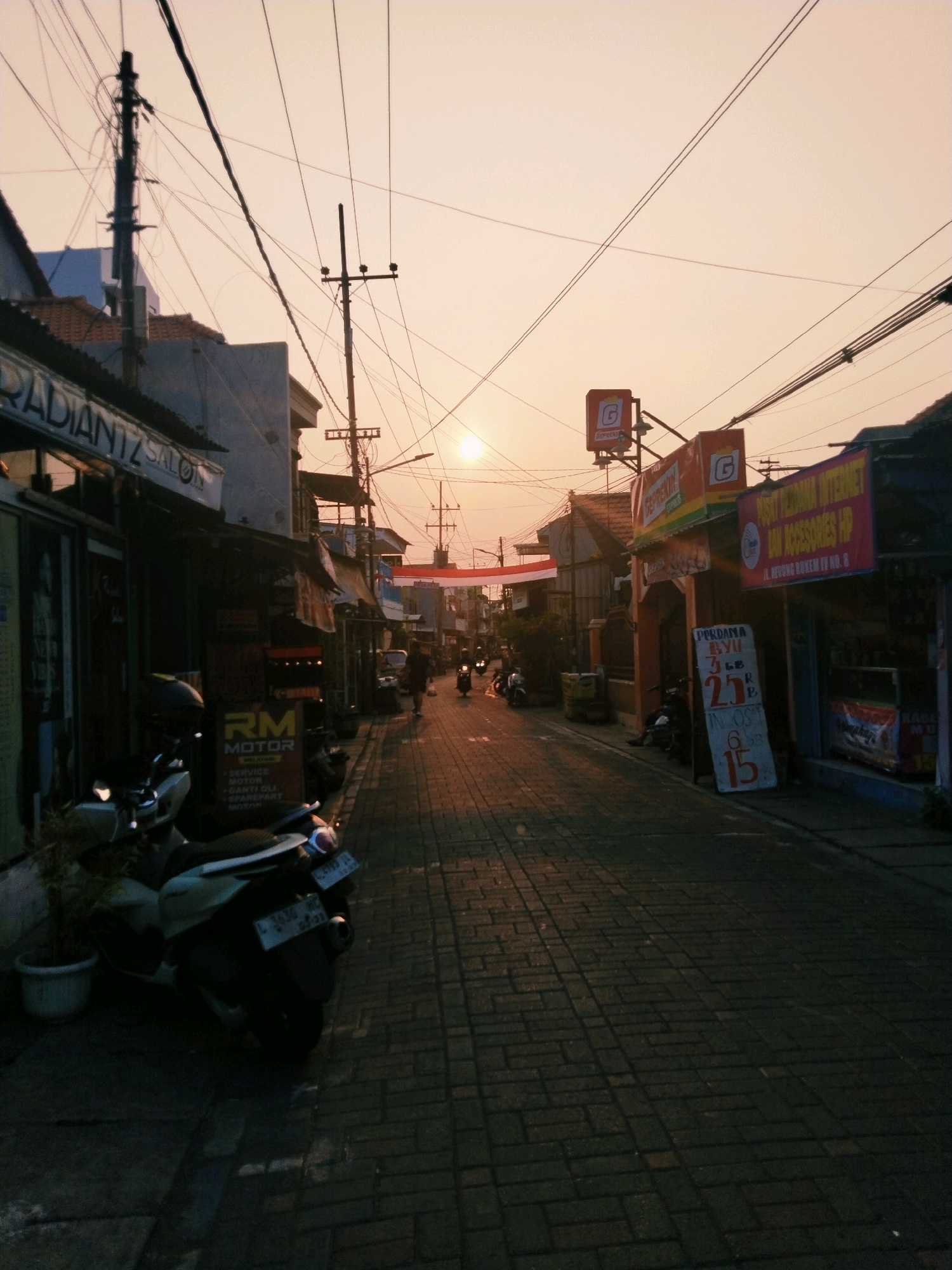 The image captures a street scene in Surabaya, Indonesia, during sunset. The sky is a warm gradient of orange and yellow, casting a soft glow over the urban environment. A narrow, paved street, likely made of brick or stone, runs diagonally from the lower left to the upper right of the frame. The street is lined with buildings on both sides, featuring storefronts with signage.
On the left side of the street, a building with a sign reading "RADIANTZ SALON" is visible. In front of it, a white scooter is parked, with a potted plant to its left. Further down the street on the left, another sign for "RM MOTOR" is displayed.
The right side of the street also has several buildings with shops. Signs are visible, including one that appears to advertise internet services and accessories. A white sign with red text, possibly listing prices, stands near the sidewalk.
Several motorcycles are parked along the sides of the street, and a few are seen moving in the distance. A person is walking down the street on the right side. The street is adorned with numerous electrical wires and utility poles, characteristic of urban infrastructure. The overall atmosphere is that of a busy, yet tranquil, late afternoon in a commercial area.