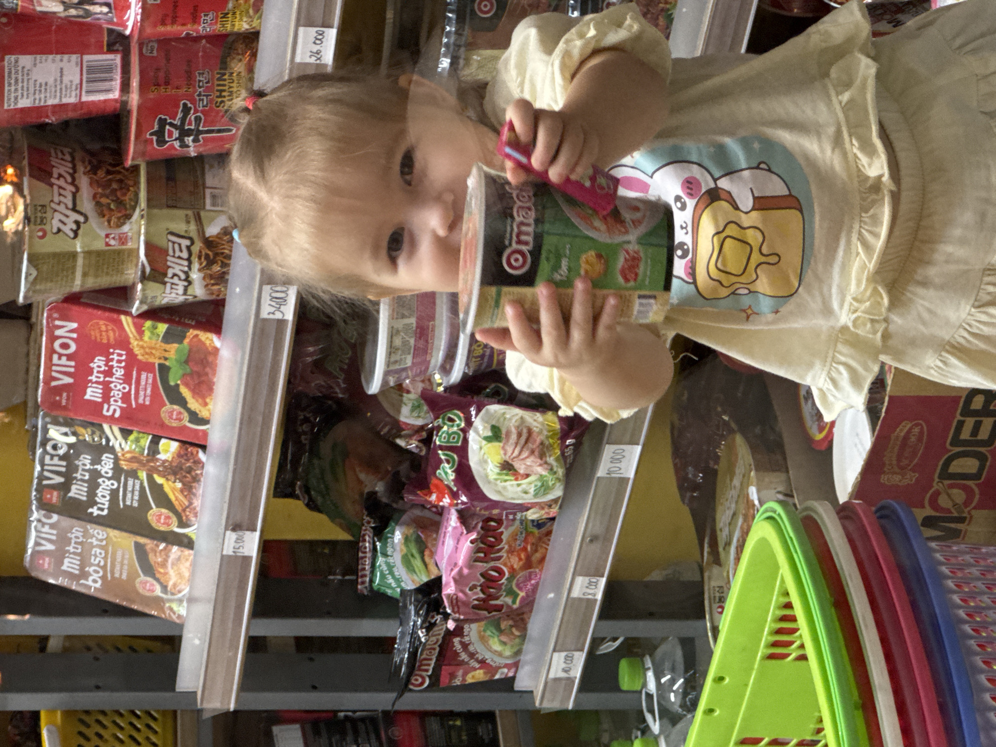 This media file shows a close-up shot of a young child, likely a toddler, holding a cup of instant noodles. The child is wearing a light yellow, ruffled shirt with a cartoon bunny design. The child's face is turned towards the cup, with an inquisitive expression. The background consists of shelves stocked with various packaged instant noodle products, indicating a retail setting, possibly a supermarket or convenience store. Many of the noodle packages are brightly colored and feature Vietnamese text and imagery.
Visible text includes brand names like "VIFON," "Hảo Hào," and "Shin Ramyun." Prices are also visible on shelf tags, such as "15.000," "34.000," "26.000," "10.000," and "8.000," suggesting the location is in Vietnam where currency is denominated in Vietnamese Dong. The lighting appears to be artificial, typical of an indoor store environment. The overall scene captures a moment of childhood curiosity and interaction with consumer goods in a Vietnamese retail space.
