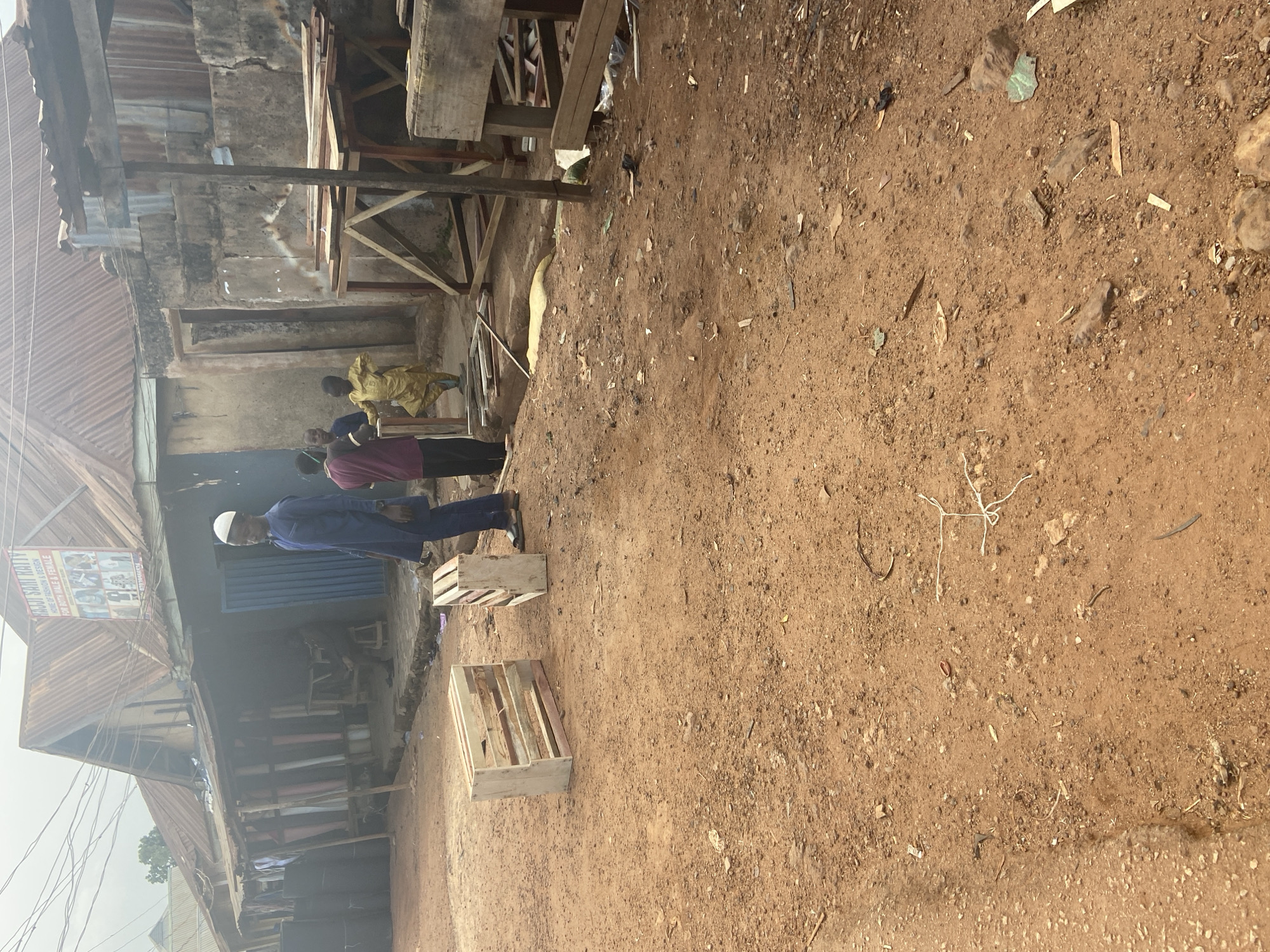 The image depicts an outdoor scene in what appears to be a market or commercial area in Bukuru, Nigeria. The ground is a reddish-brown dirt, scattered with debris.
Several individuals are present. In the foreground, a man wearing a blue robe and white cap is visible. Behind him, a group of people are gathered, one of whom is wearing a yellow garment. The figures are casually standing and interacting.
The setting includes structures that suggest shops or stalls. There are buildings with corrugated metal roofs and walls, some appearing weathered. Wooden supports and shelving are visible, indicating a makeshift or traditional market environment. Two wooden crates are placed on the ground in the foreground.
The lighting suggests it is daytime, possibly late morning or early afternoon, with shadows indicating the sun's position. The weather appears to be dry, given the dusty ground.
On one of the buildings, a sign with text and images is visible. The text appears to include "HAJJISAN KANJI", "HOUSE OF FASHION & DESIGN", and "FOR MOTHERS & FATHERS". This suggests a fashion-related business. The overall atmosphere seems to be one of everyday commerce.