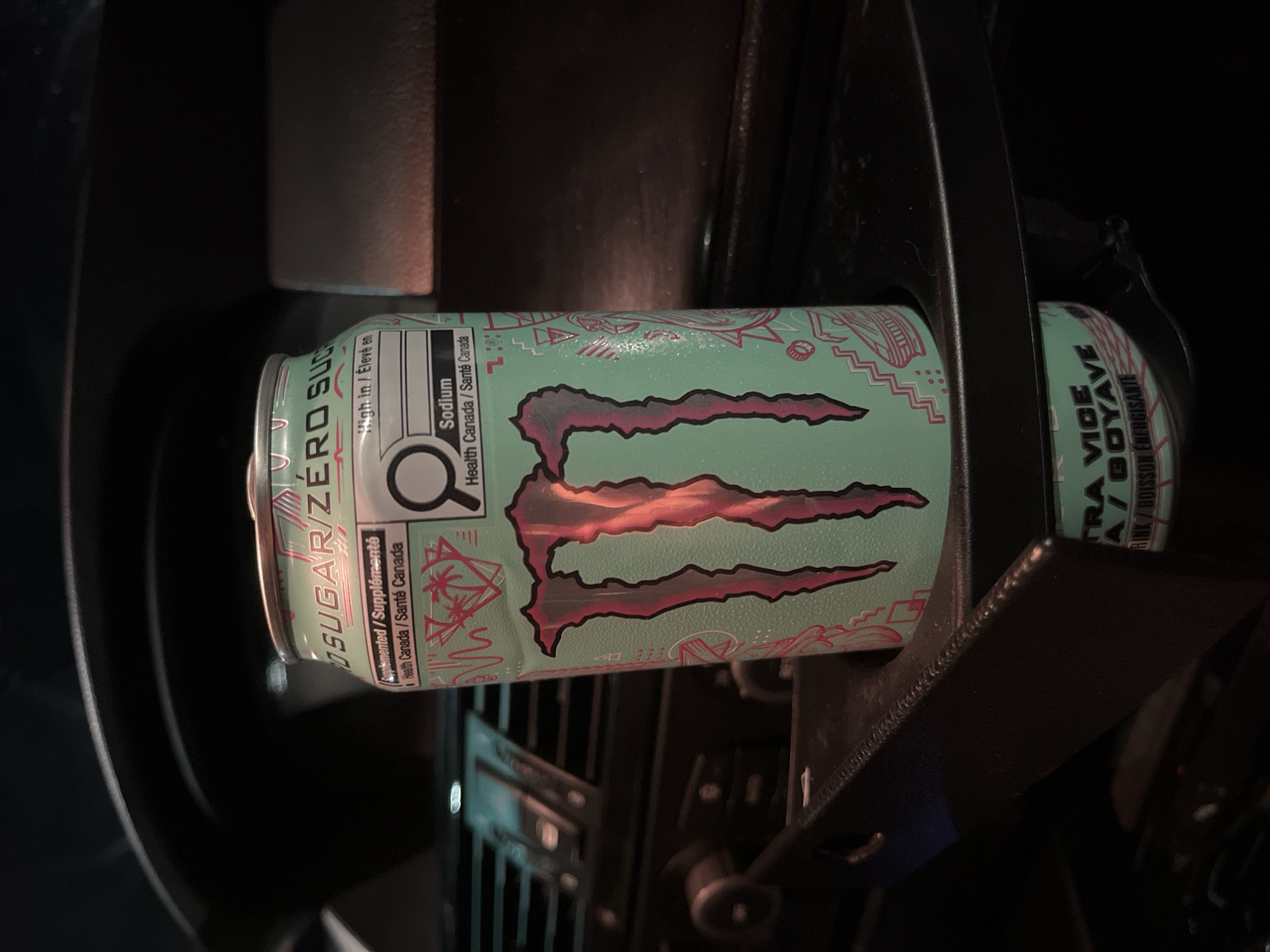 A partially visible can of Monster Zero Sugar Ultra Violet energy drink sits in an automotive cup holder. The can is light green with the Monster logo in shades of pink and red. Text on the can includes "High in/Élevé en Sodium", "Health Canada/Santé Canada", and "TRA VICE A/GOYAVE". The cup holder is black and appears to be part of the car's dashboard and interior. The scene is dark and details about the surrounding environment are obscured. The location is not Shumen, Bulgaria, but likely in a car, and the provenance of the image is unknown.