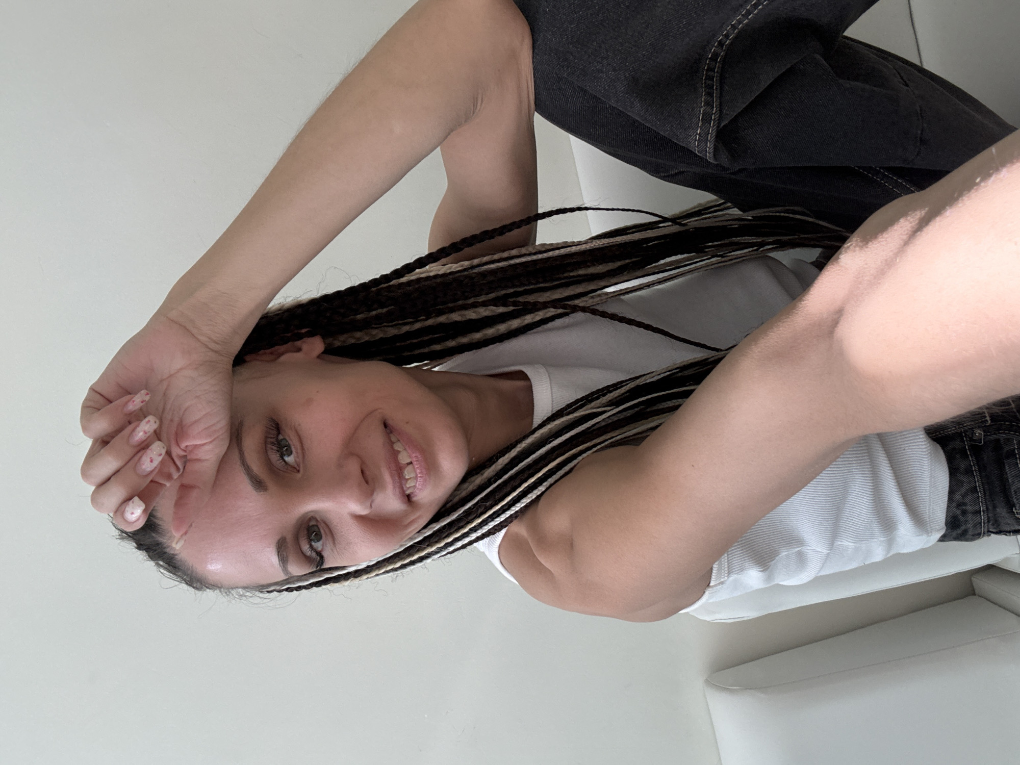 A young woman with braided hair, wearing a white t-shirt and dark jeans, is pictured indoors. She is smiling and has her hand on her forehead. She is sitting on a white couch. The location is not discernible as being in Rusanovo, Russia.