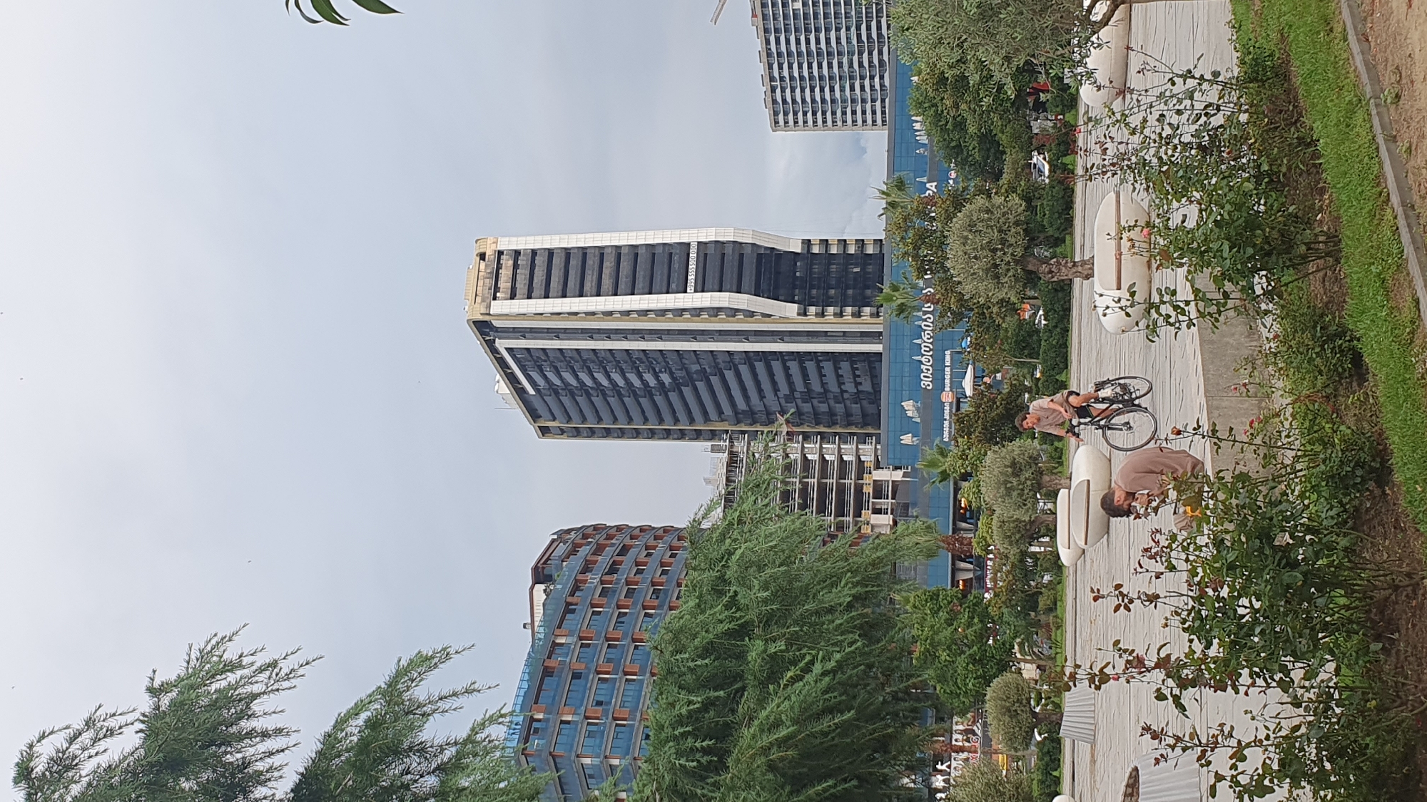 The image depicts a city scene with a prominent modern skyscraper and an adjacent curved building, both featuring extensive glass facades. In the foreground, lush green trees and rose bushes line a paved walkway. Two individuals are present: one is seated on a concrete ledge, appearing to be looking at their phone, while another person is cycling past on a bicycle. The background reveals more urban development, including a building under construction and another high-rise structure. A large blue building with visible signage, including what appears to be a "Burger King" logo, spans across the mid-ground. The sky is overcast and gray, suggesting a cloudy day. The overall setting suggests a public area within the city of Batumi, Georgia, likely a waterfront promenade or park given the presence of greenery and modern architecture.