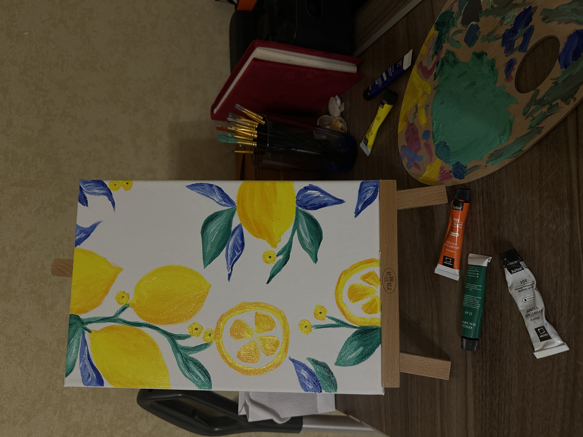 Here is a detailed description of the image:
The image shows a still life of an artist's setup. In the foreground is a canvas propped on a small easel. The canvas has a painting of lemons and leaves on it, done in yellow, green, and blue hues. Behind the canvas, various items are arranged, including a red notebook, a jar of paintbrushes, a small container with shells, tubes of paint (yellow, blue, orange, green, and white), and a wooden palette covered in blobs of dried paint.
The canvas is situated at the bottom of the frame. The other art supplies are above and slightly behind it. The background consists of a light brown, textured surface. The overall setting appears to be indoors.