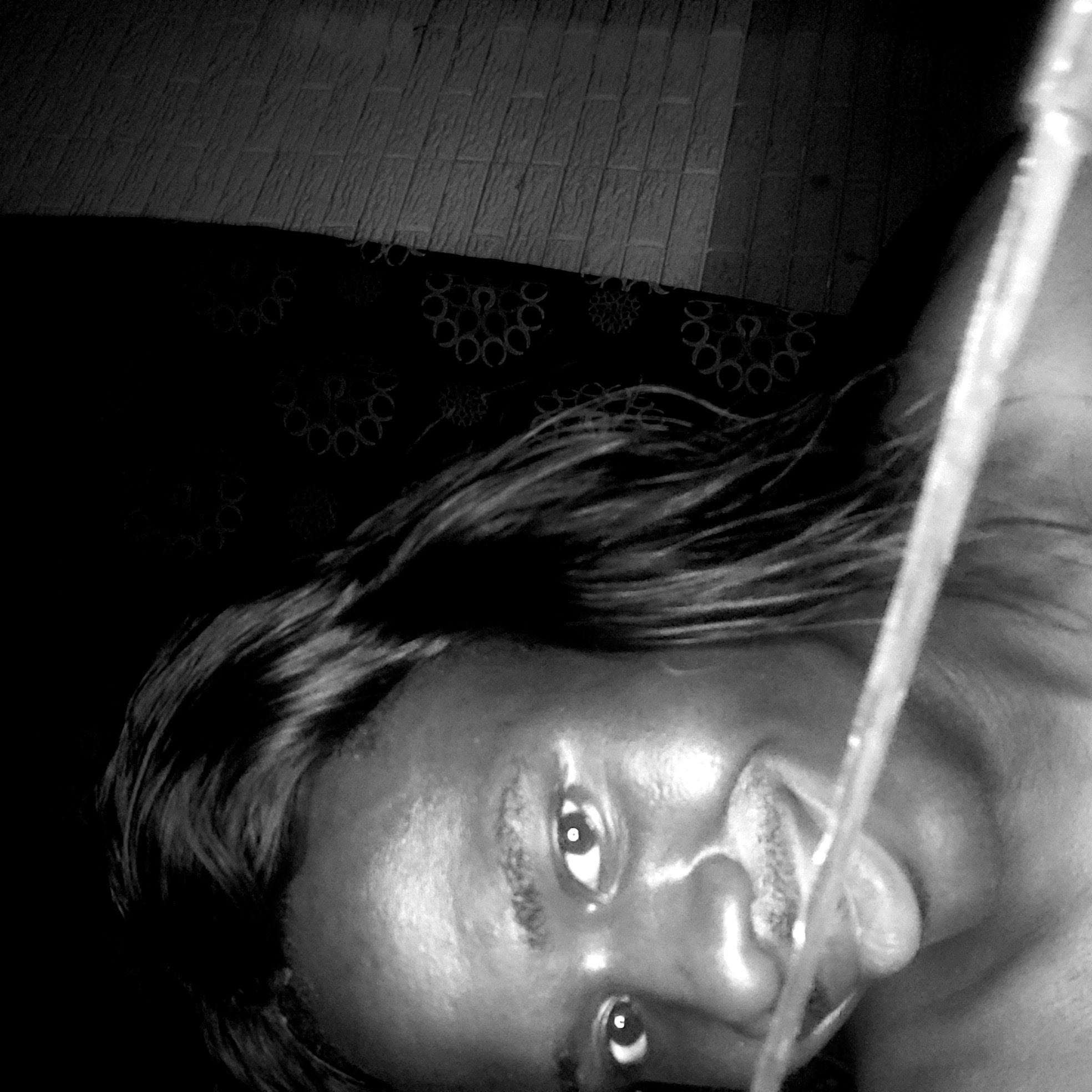 A black and white, high-angle, close-up image depicts a person lying on a surface with patterned fabric, against a wall of horizontal panels. The person has dark skin, dark hair, and is looking directly at the camera with their tongue protruding. A clear, linear object, possibly a tube or stream of liquid, is positioned vertically across their face. The location is assumed to be in Narayi, Nigeria, due to the prompt's specification.
