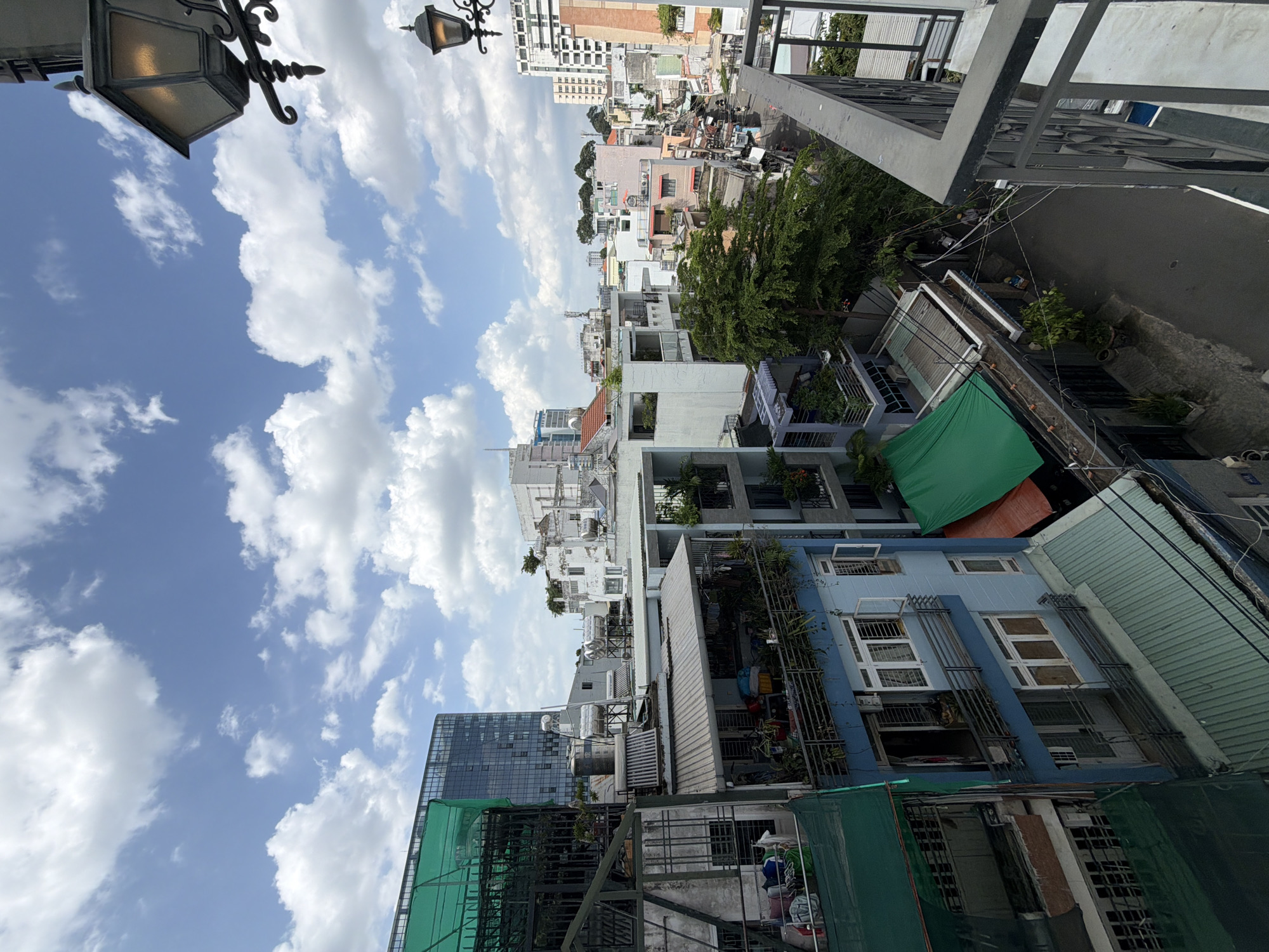 The image, rotated 90 degrees clockwise, presents a detailed view of a bustling urban environment, typical of Thủ Đức, Vietnam, seen from an elevated vantage point such as a balcony or rooftop. The scene is illuminated by bright daylight under a clear blue sky adorned with numerous fluffy white cumulus clouds.
In the rotated foreground, two ornate, dark-colored street lamps or decorative lights with textured glass shades are prominently visible. A sturdy dark metal railing, part of the observer's location, frames the right side of the shot.
The mid-ground and background showcase a dense tapestry of buildings, reflecting a mix of residential and commercial structures. These multi-story buildings display varied architectural styles, from older constructions with tiled or corrugated metal roofs to more modern designs, including a tall building with a glass facade visible towards the bottom left (when correctly oriented). Many buildings feature balconies adorned with potted plants, and some rooftops host water tanks and air conditioning units. Scattered trees and lush greenery provide pockets of natural color amidst the urban sprawl. Several green tarps or nets are visible covering sections of buildings, possibly for shade or construction. A narrow road or alleyway meanders through the central-right portion of the frame, with a network of power lines crisscrossing overhead.
The time of day appears to be late morning or early afternoon, indicated by the bright, overhead sunlight and the distinct shadows. No direct human activity is discernible, and no legible text is visible on any signs or buildings, offering a quiet, observational snapshot of a vibrant Southeast Asian cityscape.