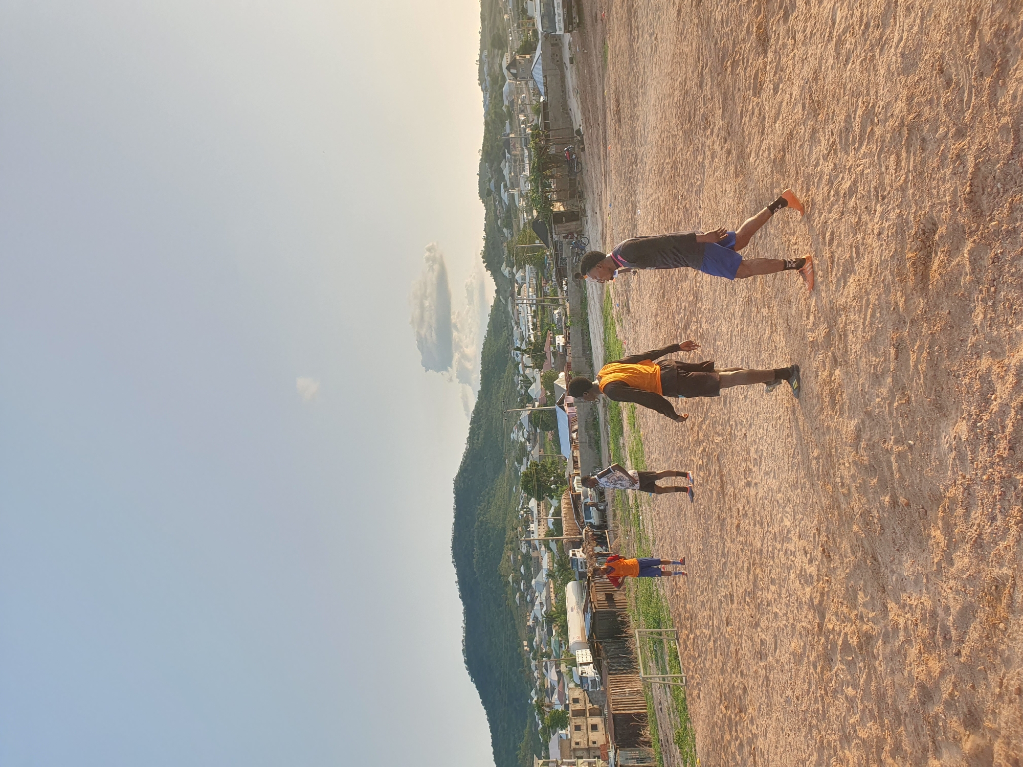 The image depicts a dirt field in Lokoja, Nigeria. Four people are present, appearing to be engaged in physical activity. The first person is in the top right of the image, with a dark shirt, blue shorts, and orange shoes, and appears to be stretching or balancing. The second person has a dark shirt with an orange bib, and dark colored shoes. The third person wears a white patterned shirt and dark shorts, and the fourth person is in an orange shirt with dark shorts. A small, simple goal post is in the lower portion of the image. Behind the field, a town with a mixture of buildings is visible, backed by a tree-covered mountain. The sky is light blue with some scattered clouds.