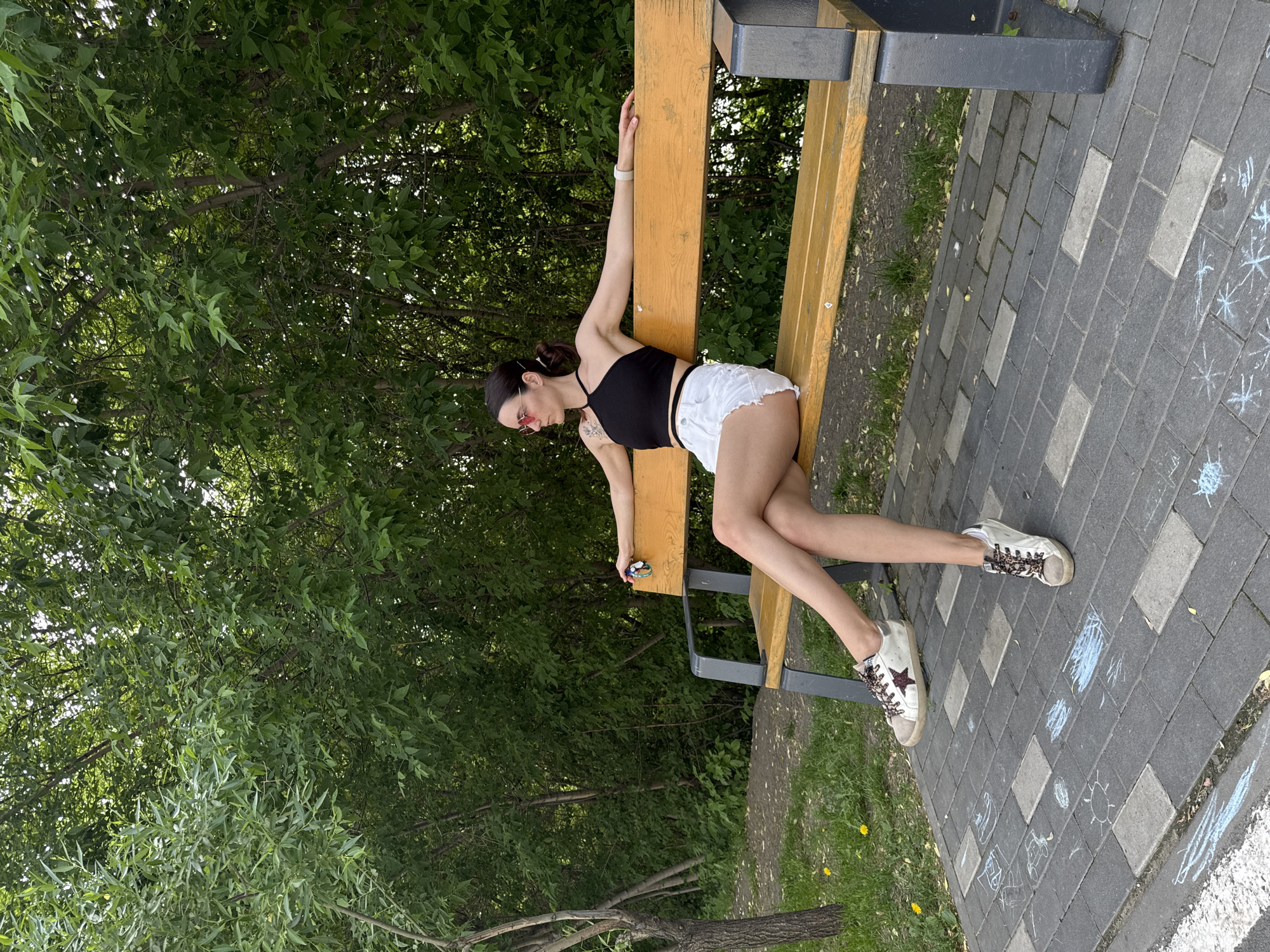 A young woman is reclining on a bench in what appears to be a park, possibly located in Tyumen, Russia, based on visual cues. She is wearing a black top, white shorts, and sneakers, and she has one arm extended above her head towards the back of the bench. In her other hand, she is holding a small object, perhaps a phone or a similar device. The bench is made of wood and metal, and it is situated on a paved area with brick patterns. The pavement shows some chalk drawings. The background consists of green trees and foliage, suggesting a natural or landscaped setting.