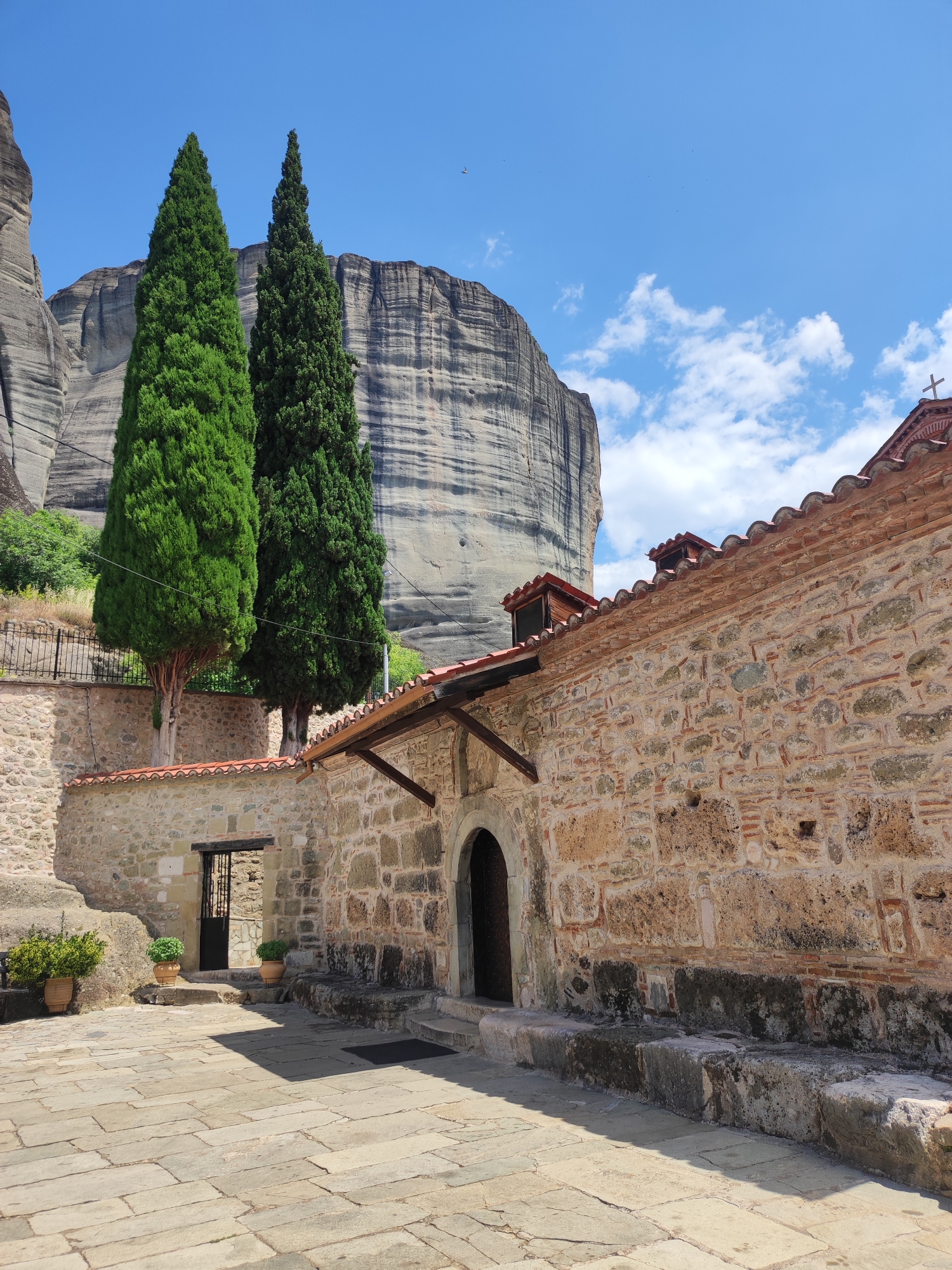 A full, eye-level, outdoor, long-shot captures a stone-paved courtyard leading to an ancient monastery. Two tall, slender, green cypress trees stand in front of a massive, weathered rock formation. The monastery building is constructed of rough-hewn stones and features a red-tiled roof. An open doorway with an arched entrance reveals a dark interior. In the background, a portion of a rocky cliff face rises majestically under a bright blue sky dotted with white clouds. The scene is bathed in daylight, casting shadows across the courtyard. The location is the monastery of Meteora in Kalabaka, Greece.