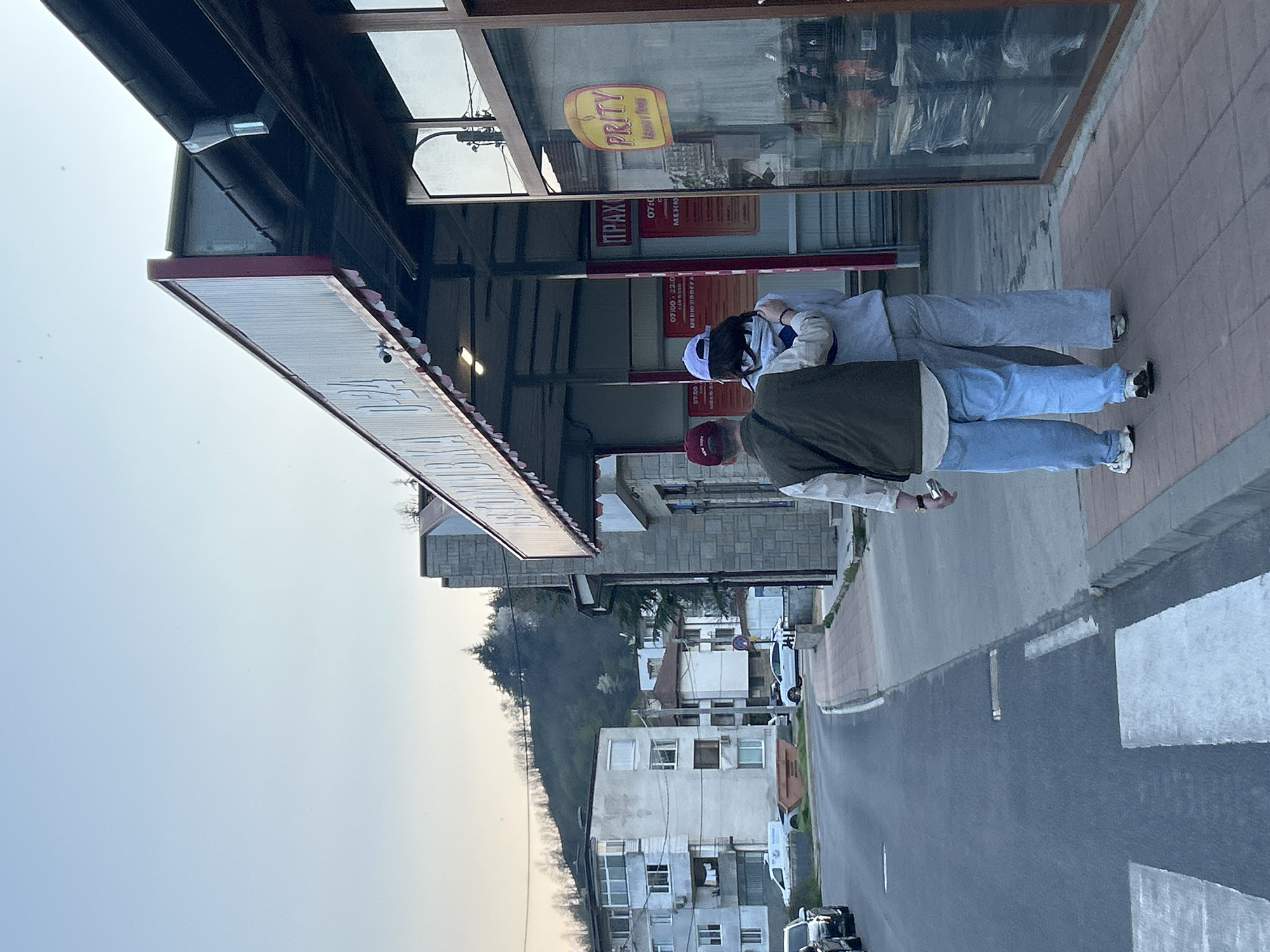 The image depicts a street scene in Zlatograd, Bulgaria, featuring two individuals standing outside a shop. The shop has a prominent sign that reads "PRITY" and "ПРАХ". One person wears a red baseball cap, a green jacket, and blue jeans, and is holding a silver object in their hand. The other person wears a white cap and blue athletic clothing. The street runs through a residential area with buildings of varying heights and architectural styles, and several cars are parked along the road. The sky is visible with diffused light, suggesting either early morning or late afternoon. A zebra crossing is visible on the street.