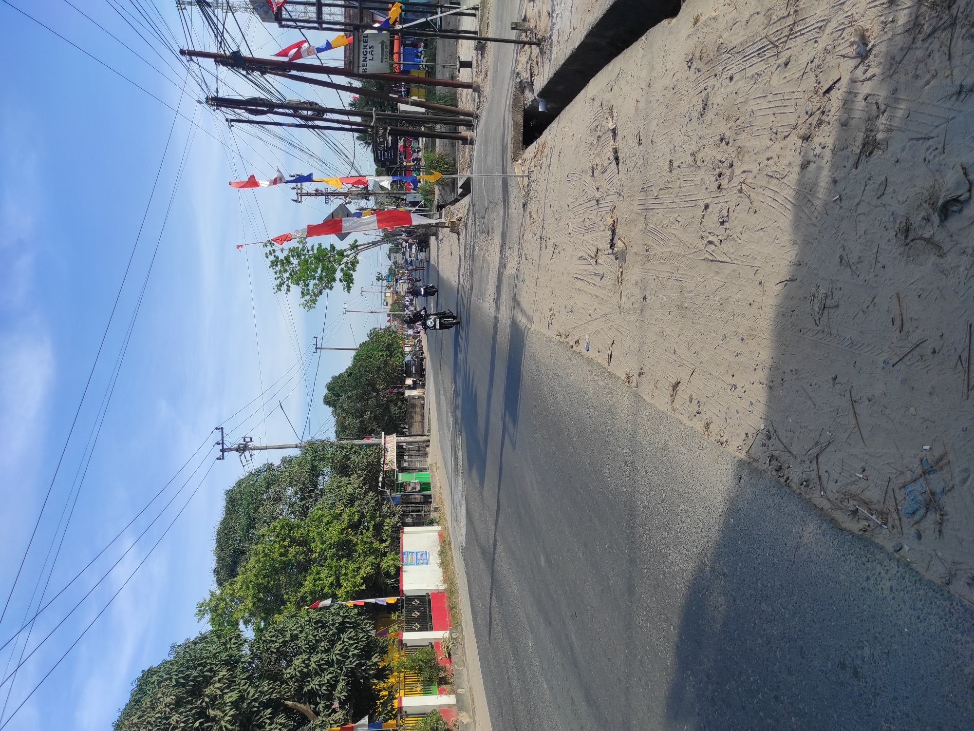 The image depicts a street scene in Muara Jawa Ulu, Indonesia, under a clear blue sky. The street is paved with asphalt and runs from the foreground towards the background, with a sandy, untidy sidewalk on the right side. Several red and white Indonesian flags are prominently displayed along the roadside, some on poles and others attached to utility poles. A line of trees with dense green foliage borders the left side of the street, behind a low wall.

In the street, several motorcycles and a tricycle are in motion. On the left side of the street, buildings and a fence with yellow and red accents are visible. On the right side, utility poles with numerous wires are clustered together. A sign that reads "BENGKEL LAS" (Welding Workshop) is also visible. The overall impression is of a moderately busy street in a tropical urban or semi-urban setting, likely during the daytime given the bright sunlight and shadows. The sandy area on the right shows tire tracks, suggesting vehicular traffic has passed through. A concrete drain runs along the edge of the sidewalk.