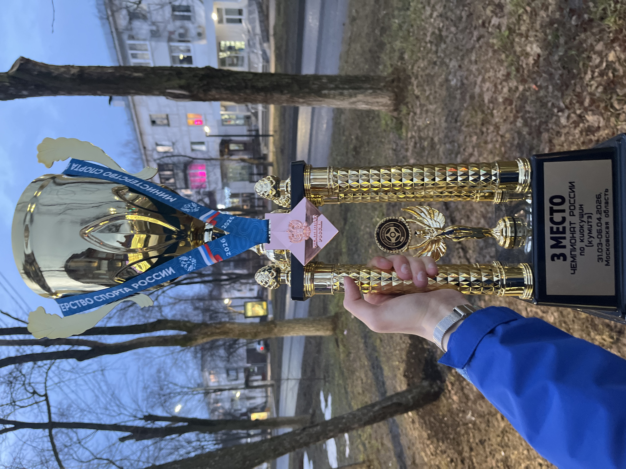 A person in a blue jacket is holding a golden trophy outdoors, presumably in the city of Chaikovsky, Russia. The trophy has a blue ribbon with Russian writing on it, and a pink emblem on top. The background shows buildings, trees, and what appears to be a street. The lighting suggests it is either early morning or late evening.