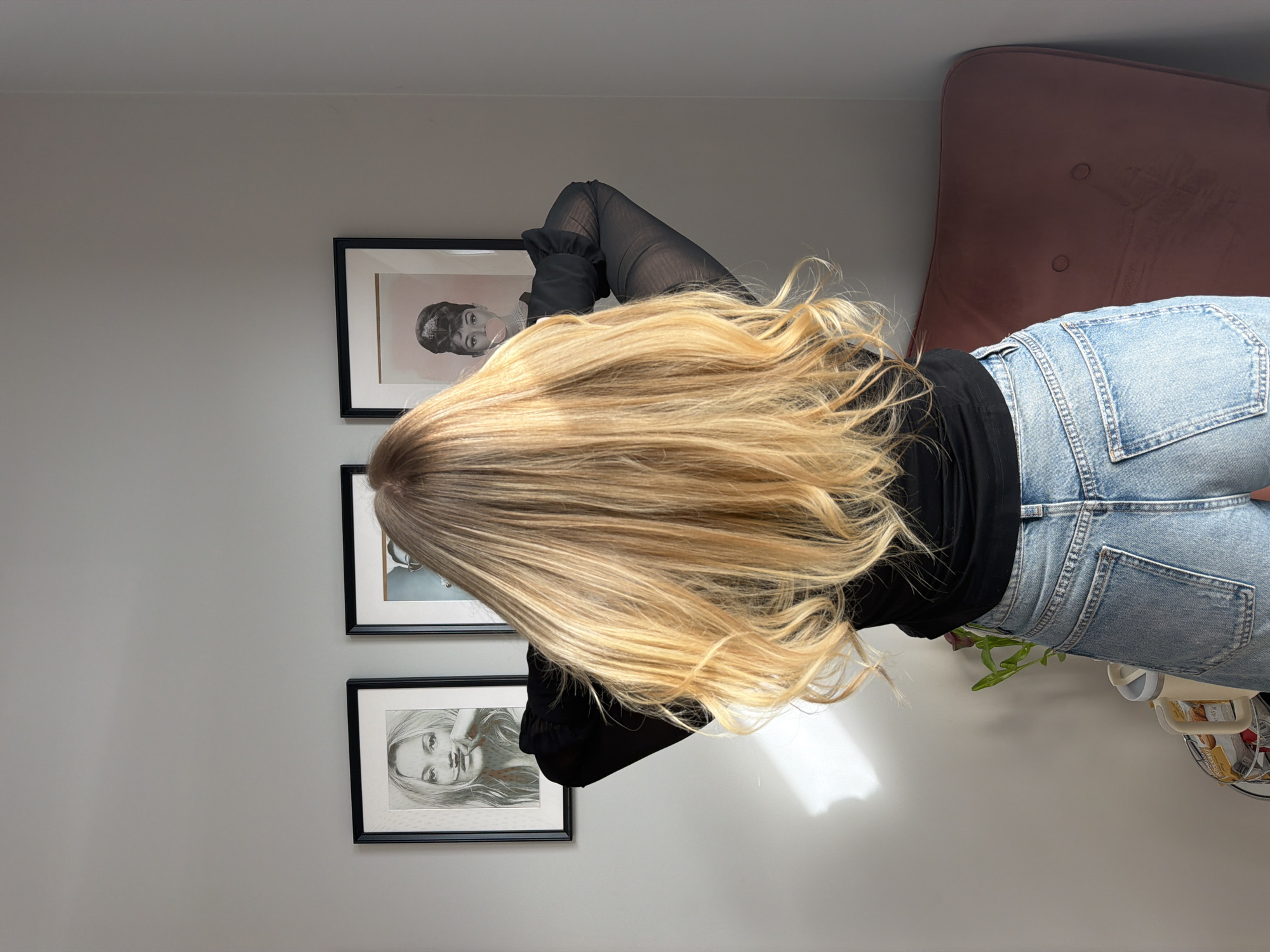 A woman with long, blonde hair is photographed from behind, facing a light grey wall adorned with three framed portraits of women. She is wearing a black top with sheer sleeves and light blue jeans. A brown chair is situated to her right. The photograph appears to be taken indoors, possibly in a home. There is no information to suggest the photograph was taken in Riga, Latvia.