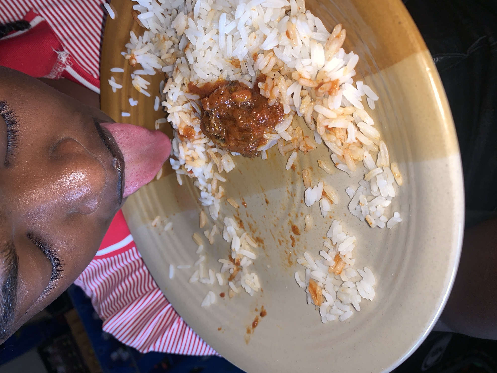 A close-up image shows the left side of a person's face in the foreground, with their tongue extended towards a plate of food. The person has dark skin, with visible long eyelashes and closed eyes, suggesting a moment of intense focus or enjoyment.
The main subject is a round, two-toned plate, primarily light beige with a brown rim, holding white rice mixed with a reddish-brown sauce or stew. A more concentrated blob of the reddish stew, possibly containing small pieces of meat or vegetables, is visible near the center of the plate. Scattered grains of rice and streaks of sauce indicate that the meal has been partially consumed or is in the process of being eaten.
The activity depicted is the person licking the plate, a common gesture of savoring the last remnants of a meal. The setting appears to be indoors, with a red and white striped fabric visible in the background, possibly part of clothing or a table covering. The lighting suggests an indoor environment. There is no visible text in the image. This scene is contextualized within New Stadium, Nigeria.