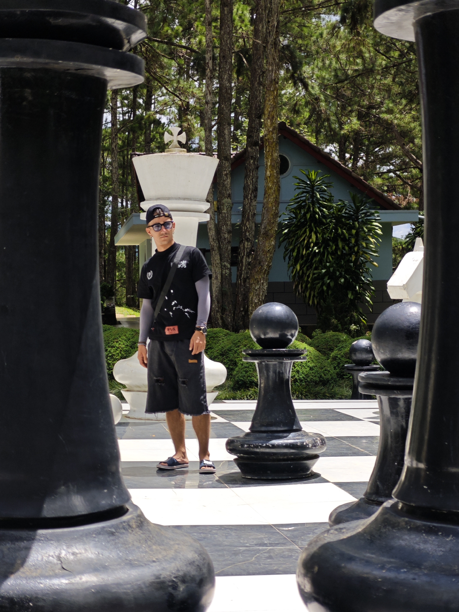 A man stands on a giant chessboard in Lâm Viên - Đà Lạt, Vietnam, a tourist destination. He is wearing a black t-shirt, dark shorts, sunglasses, and a cap. Behind him, a light blue building with a red roof is visible among pine trees. The chessboard is surrounded by lush greenery and other oversized chess pieces, including black pawns and white pieces. The man is positioned between large black chess pieces, framing him within the scene.