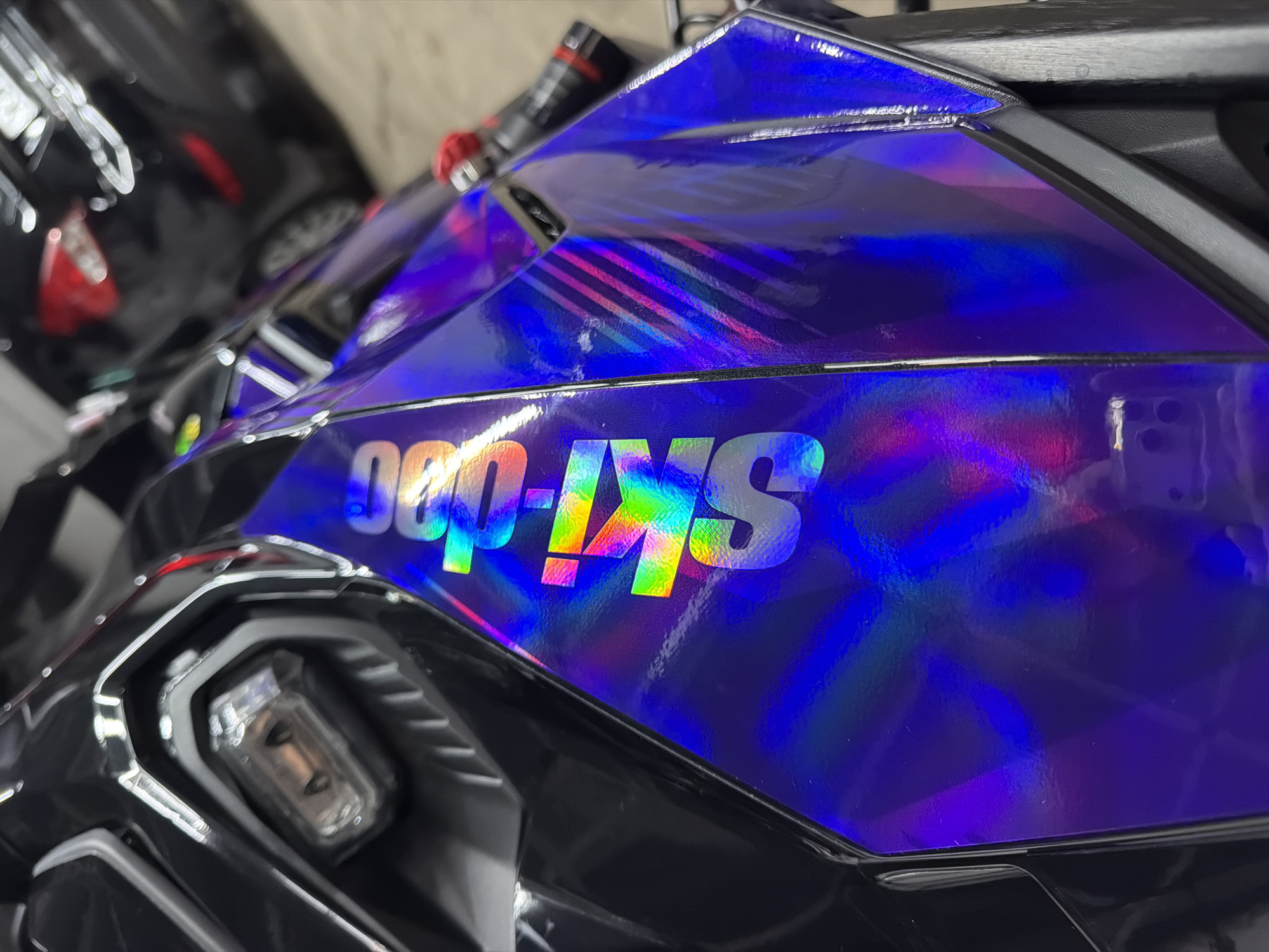 The image shows a close-up of a vehicle, likely a scooter or small motorcycle, with a glossy, holographic purple finish. "SKI-DOO" is prominently displayed in large, iridescent lettering on the vehicle's body panel. The vehicle features a black headlight assembly with a visible light element inside. In the background, part of another vehicle is visible, suggesting the scene is possibly in a garage or parking area. The location in Krasnoyarsk, Russia, is not evident in the image.