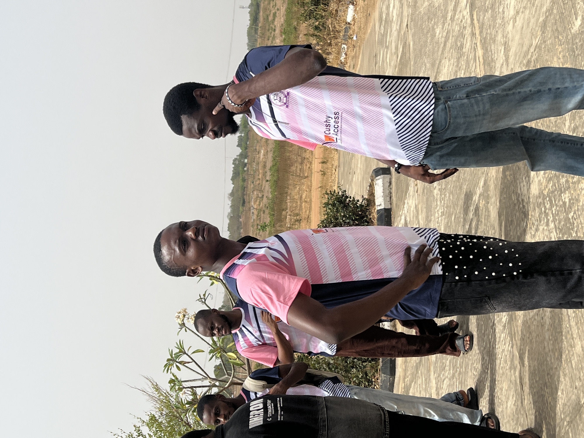 A group of at least five young men are standing outdoors on a paved surface in what appears to be a vacant or undeveloped area. The location is unspecified, but given the prompt instruction, it's likely to be in the city of Minna, Nigeria. The men are dressed in casual attire. Some are wearing matching t-shirts with pink and white stripes and the words "Cushy Access." The man at the top is wearing jeans. The environment includes some sparse vegetation and what appears to be undeveloped land in the background. The lighting suggests it is daytime. One man is touching his chin in a pensive manner, while others are interacting with each other. The overall scene appears informal and potentially candid.