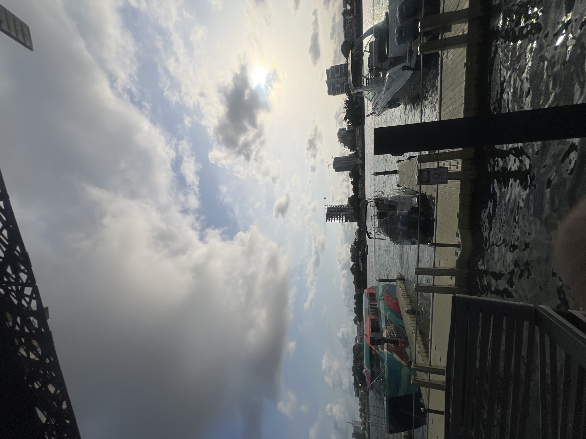 This is a photograph taken in Ikoyi, Nigeria, showing a waterfront scene with docked boats and distant city buildings. The sky is partly cloudy with the sun obscured by clouds. In the foreground, part of a metal structure, possibly a bridge or building framework, is visible on the left, and sections of a metal railing or fence are on the right. Several boats of varying sizes are docked along a wooden pier or platform. In the distance, buildings are visible on the opposite side of the water. The water surface is textured with ripples, reflecting light and the surrounding environment.