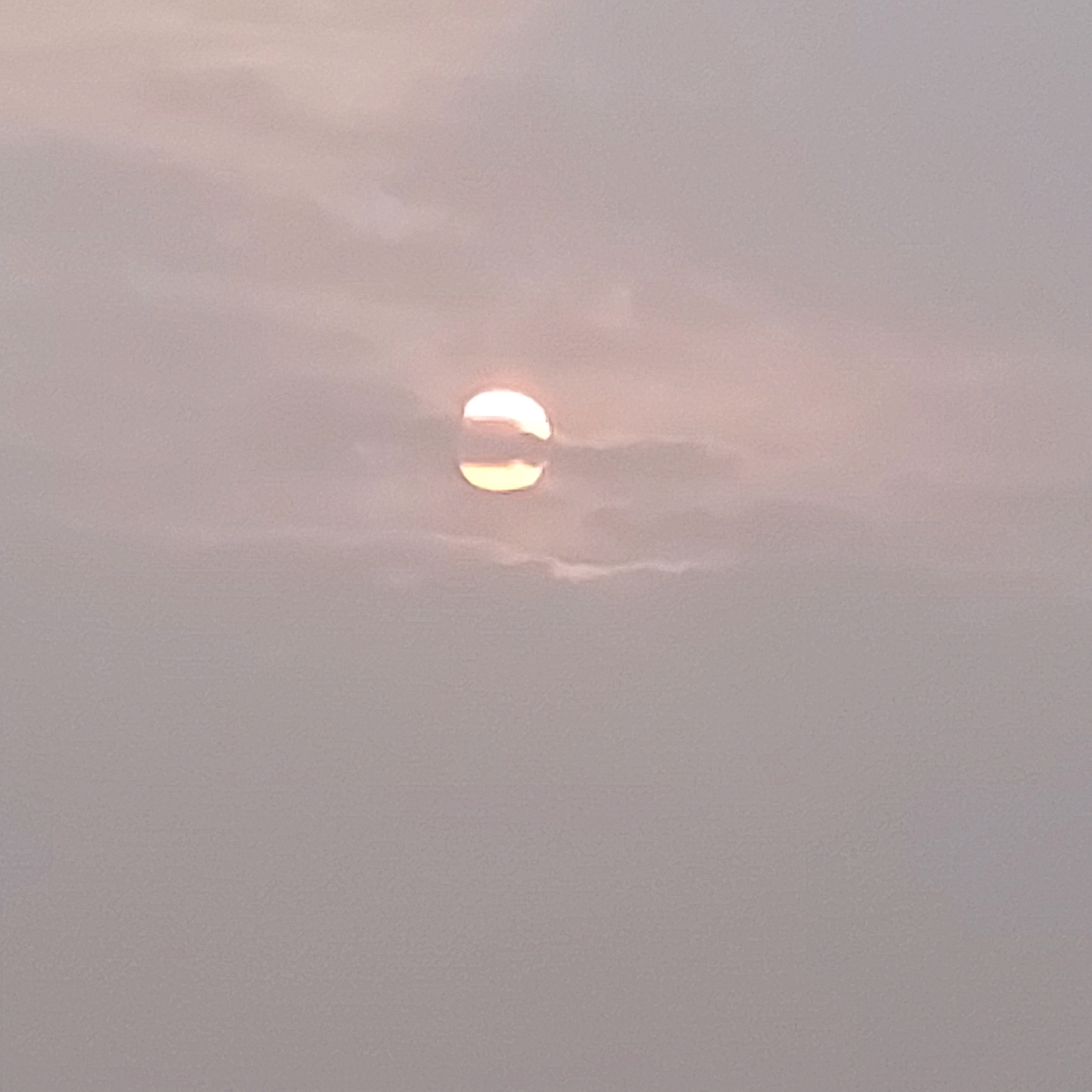 The image shows a pale, hazy sky with the sun partially obscured by clouds. The sun appears as a bright, circular shape with a horizontal band of darker cloud passing through its center, creating a visual effect similar to a yin and yang symbol. The overall color palette is muted, with shades of grey, white, and a hint of pale orange from the sun. The texture of the image suggests a slightly grainy photograph. There are no discernible human figures, objects, or specific environmental features that would indicate a particular city or location.