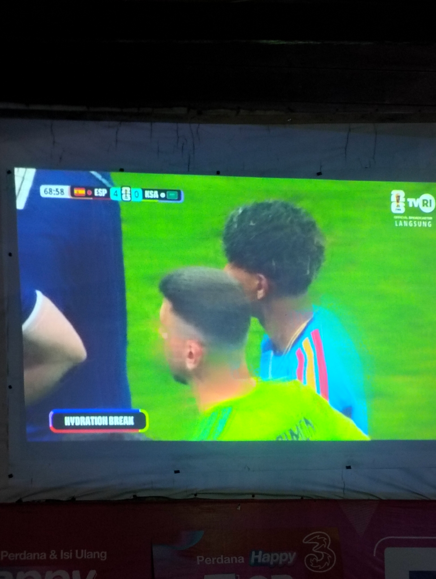 The image captures a television screen displaying a soccer match. The score is visible at the top left: 68:58, with Spain (ESP) leading Saudi Arabia (KSA) 4-0. Two players are partially visible on screen. The player on the left is wearing a dark blue jersey, and the player on the right is wearing a bright blue jersey with red and yellow stripes. In the lower middle of the screen, a notification reads "HYDRATION BREAK" within a colorful border. The television screen is projected onto a white surface, likely a screen or wall, which appears to be slightly uneven and textured. Below the projected screen, a dark red banner with text and a logo is visible, possibly indicating advertising or a channel identifier. The overall lighting suggests the image was taken in a dimly lit environment.