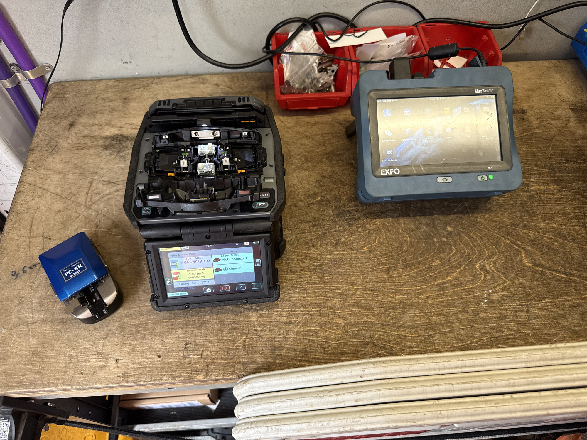 The image shows a close-up, overhead view of a wooden workbench. The workbench is cluttered with various tools and equipment related to fiber optic splicing.
In the top left quadrant of the image, a blue fiber optic cleaver is visible. To its right, a large, open fiber optic splicing machine is positioned. The machine has its internal components exposed, revealing delicate machinery and wires. Its screen displays text and graphics related to fiber optic splicing parameters.
Below the splicing machine, a red plastic container holds small metal parts and tools. A black cable snakes from the splicing machine towards the right side of the image, connecting to an unseen power source.
In the bottom right quadrant, a blue handheld device, identified by the "EXFO" logo as a MaxTester, is prominent. Its screen displays a touch interface with various icons and menus, suggesting it's in an active state, possibly performing diagnostics or settings configuration. A green indicator light is visible on the front of the device. Another black cable connects to this device, similar to the one connected to the splicing machine.
To the left of the workbench, a stack of white, coiled material, likely fiber optic cable spools, is partially visible. The background of the image shows a light-colored wall and some purple pipes secured with metal brackets.
The lighting in the scene is adequate, casting soft shadows and highlighting the details of the equipment. The overall impression is that of a workspace actively being used for fiber optic installation or maintenance.
The scene described is not located in the city of Watford, United Kingdom.