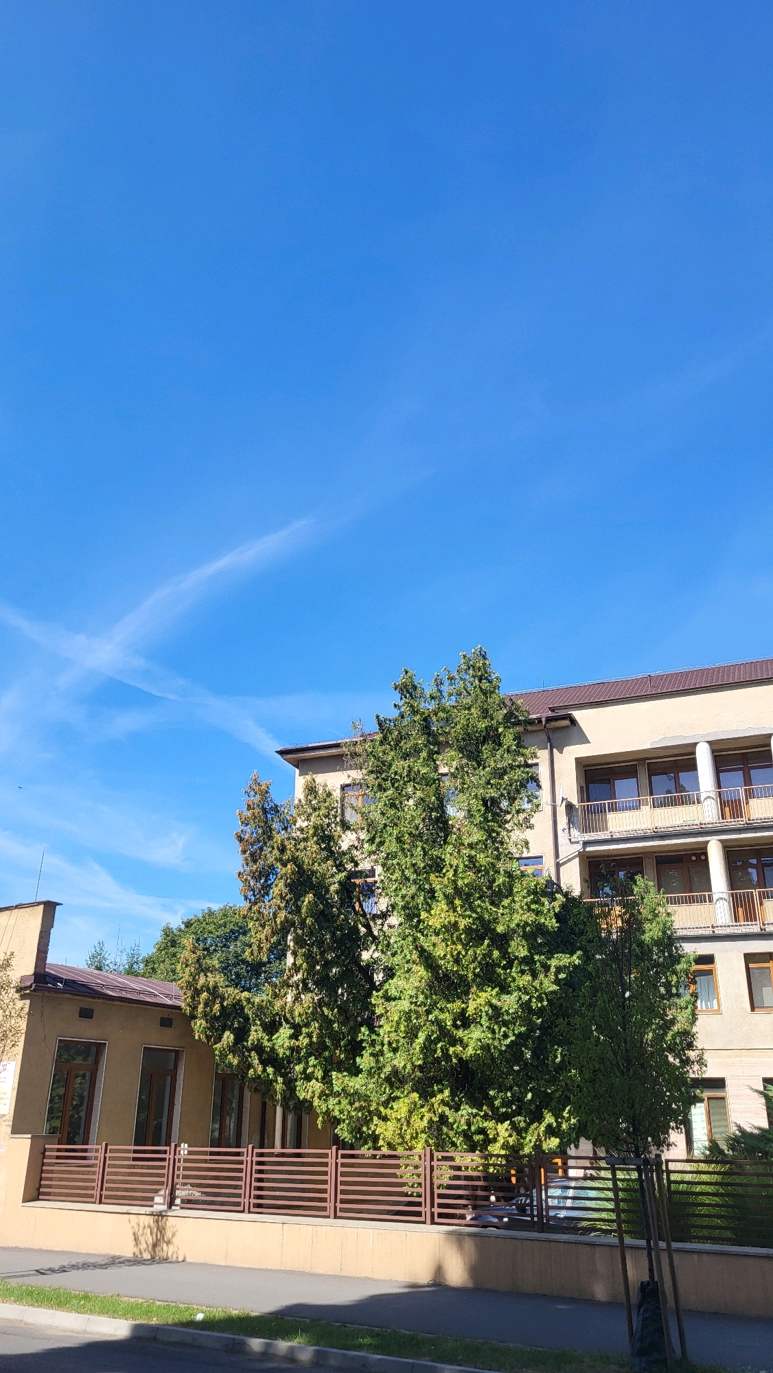 The image shows a building in Prešov, Slovakia. The building appears to be a modern, multi-story structure with a beige facade and balconies. In front of the building is a fence and some trees. The sky is clear and blue with some faint clouds. There is a street in the foreground with a sidewalk. The overall scene suggests a residential or office building in an urban setting.